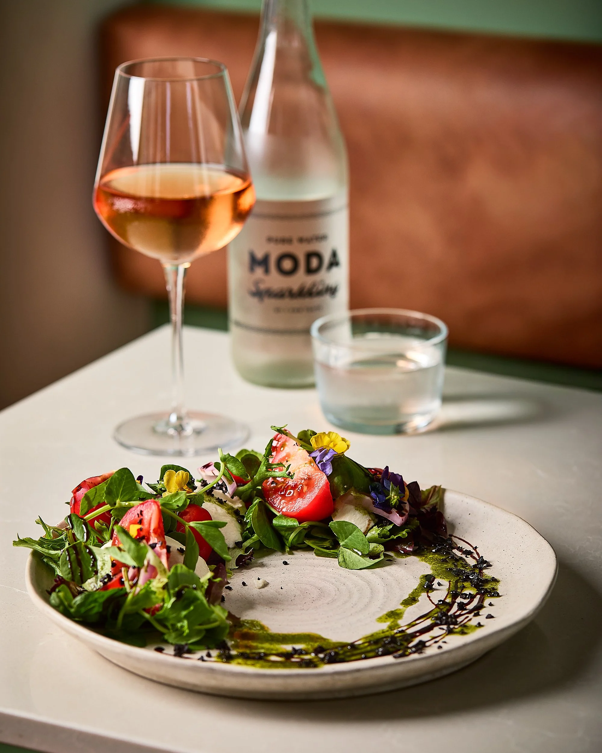 A restaurant table with a salad on a ceramic plate, a glass of rosé wine, a glass of water, and a bottle of sparkling mineral water in the background. Sydney restaurant photographer, Cafe brand photographer. 