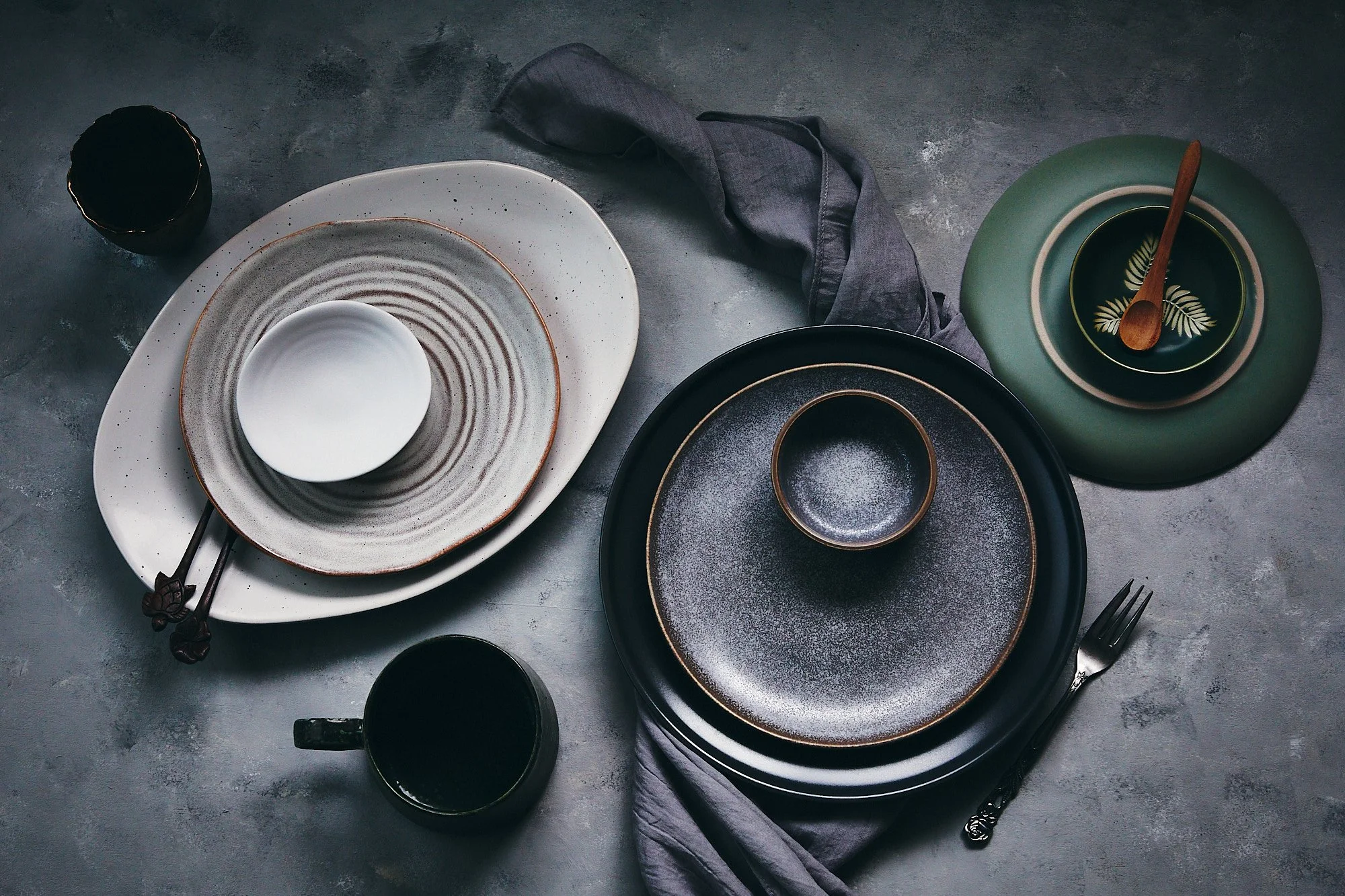 A set of empty plates, bowls, a cup, a fork, and a spoon arranged on a grey surface, with a gray napkin and some drinking cups nearby. Commercial photography of products, lifestyle photo