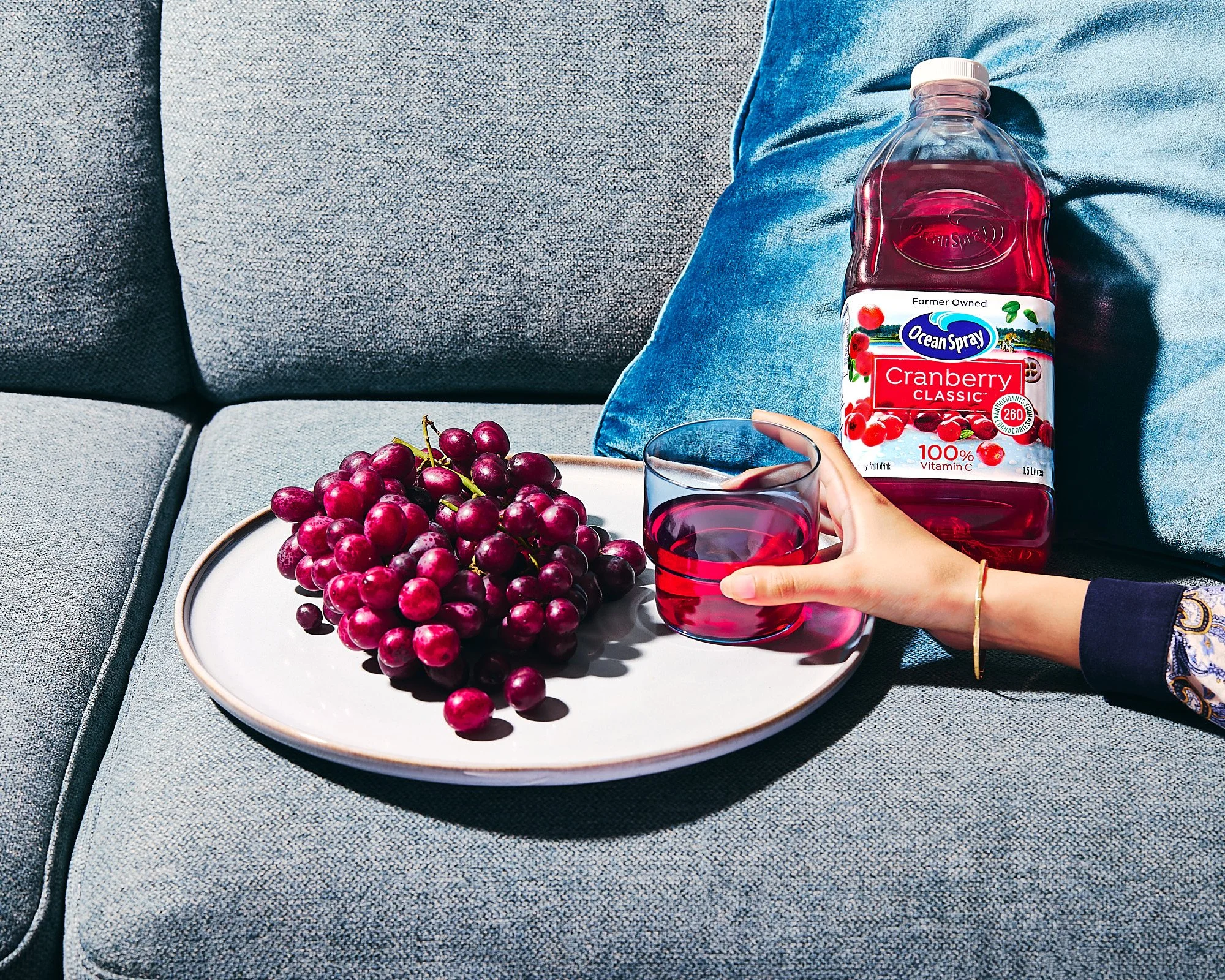 A person holding a glass of cranberry juice on a gray upholstered sofa next to a white plate of red grapes and a bottle of Ocean Spray cranberry juice. Advertising photography in Sydney, product photographer 