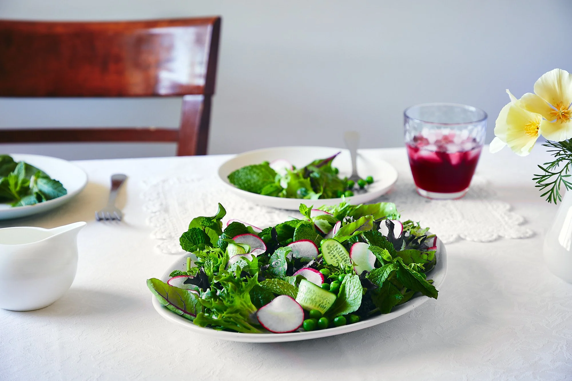 Fresh green salad with radishes, cucumbers, and peas on a white plate on a dining table with a glass of red beverage, a small white pitcher, and a vase with pale yellow flowers. Bright food photography