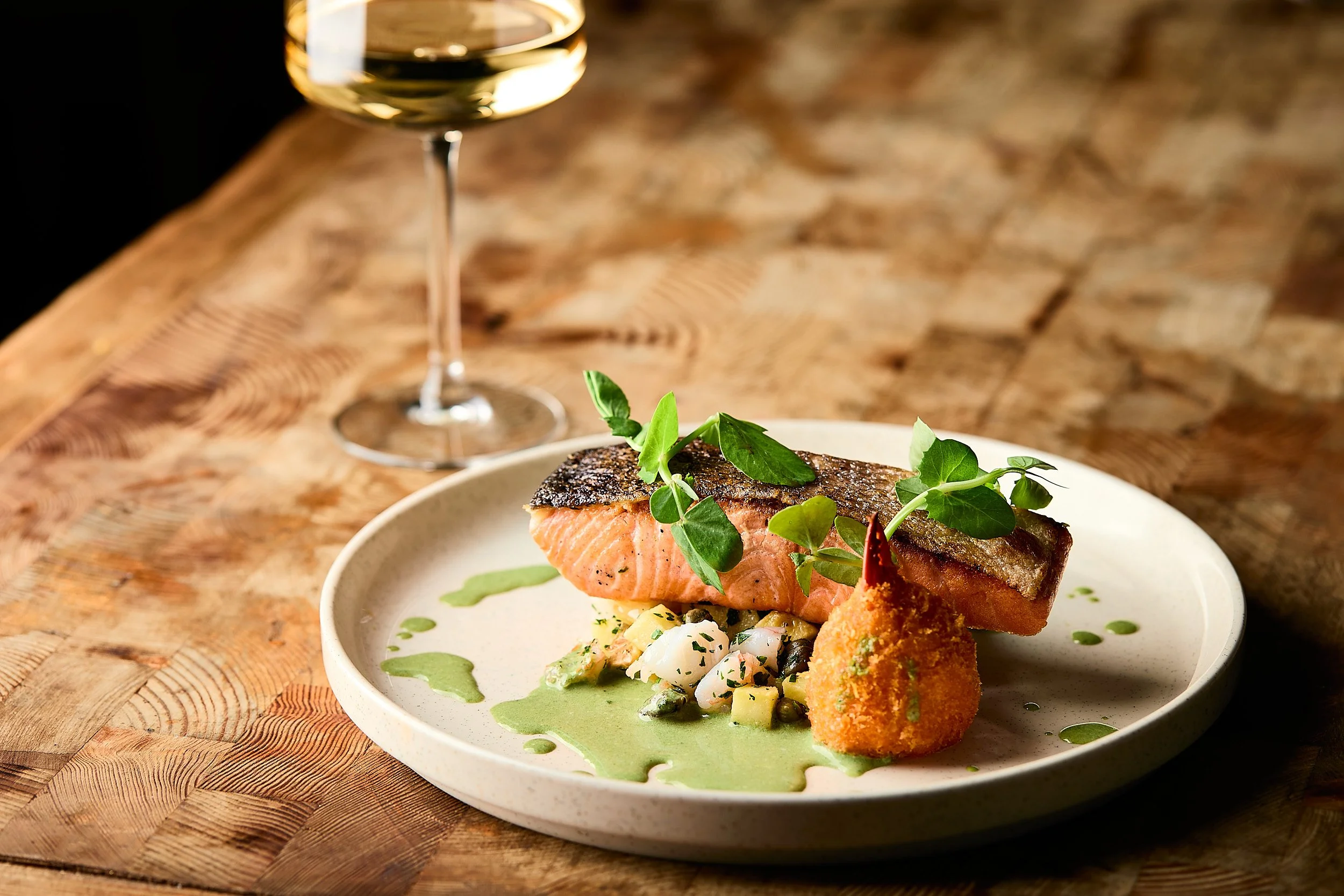 A plated dish of grilled salmon with herbs, accompanied by a green sauce, garnished with fresh herbs, served on a white plate with a glass of white wine in the background on a wooden table. Sydney Restaurant photographer, brand photographer.   