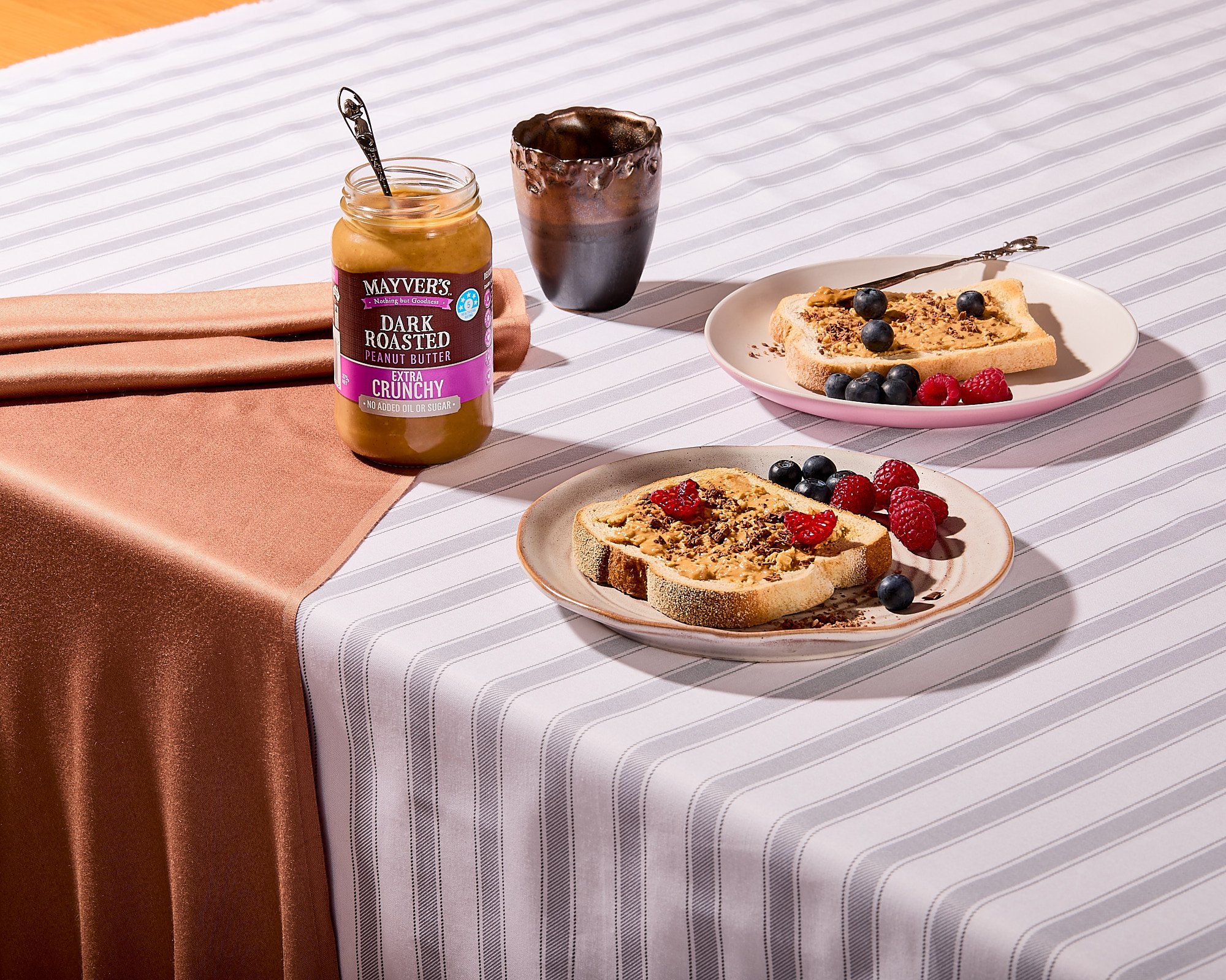 Table set with two plates of toast topped with peanut butter, berries, and chocolate shavings, a jar of peanut butter, a cup of coffee, and a pink cloth on a striped tablecloth. Product photography, brand photographer. Photo taken by Mah Vafaei.