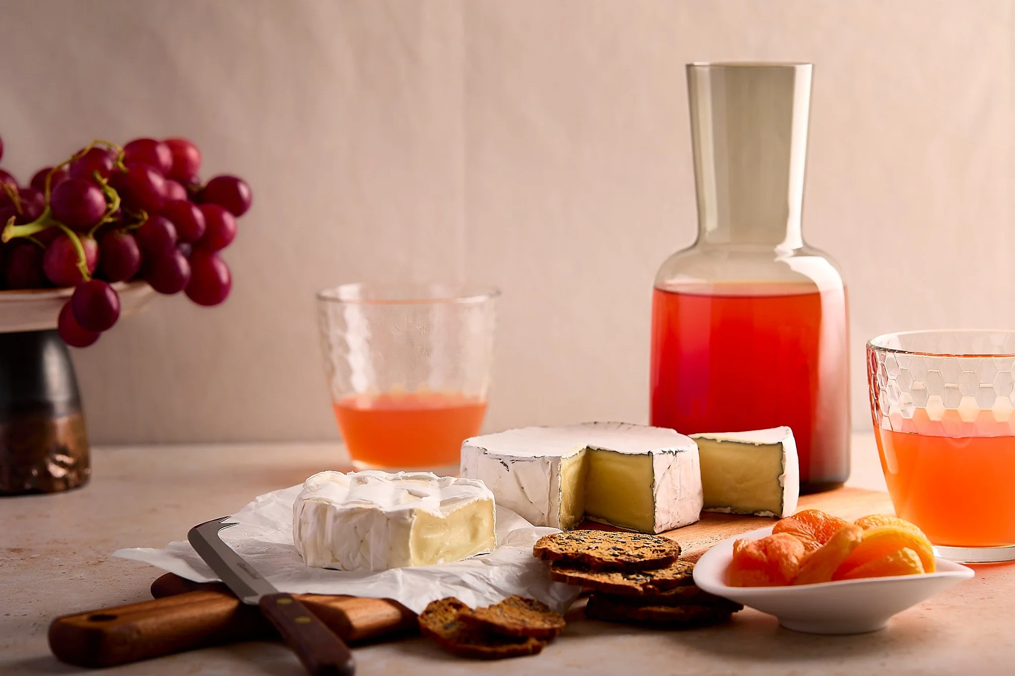A cheese platter with brie cheese, fruit preserves, and sliced fruit, accompanied by two glasses of pink beverage, a bottle of pink liquid, and a bunch of red grapes in the background. High-end food photography.