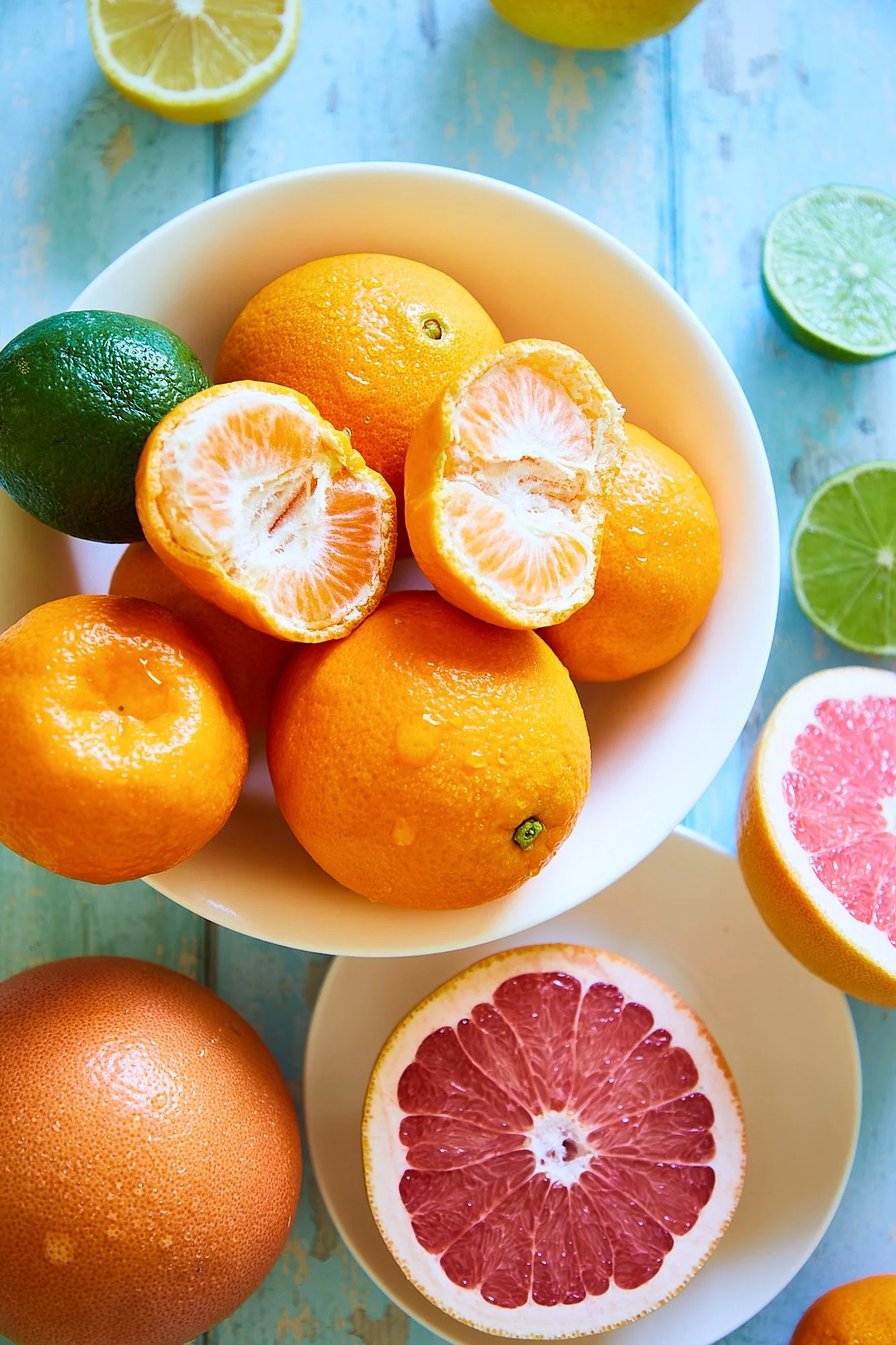 A bowl with lemons, oranges, and a lime on a blue wooden surface, with additional citrus slices on the table. Commercial photography for super markets, fresh food photography.