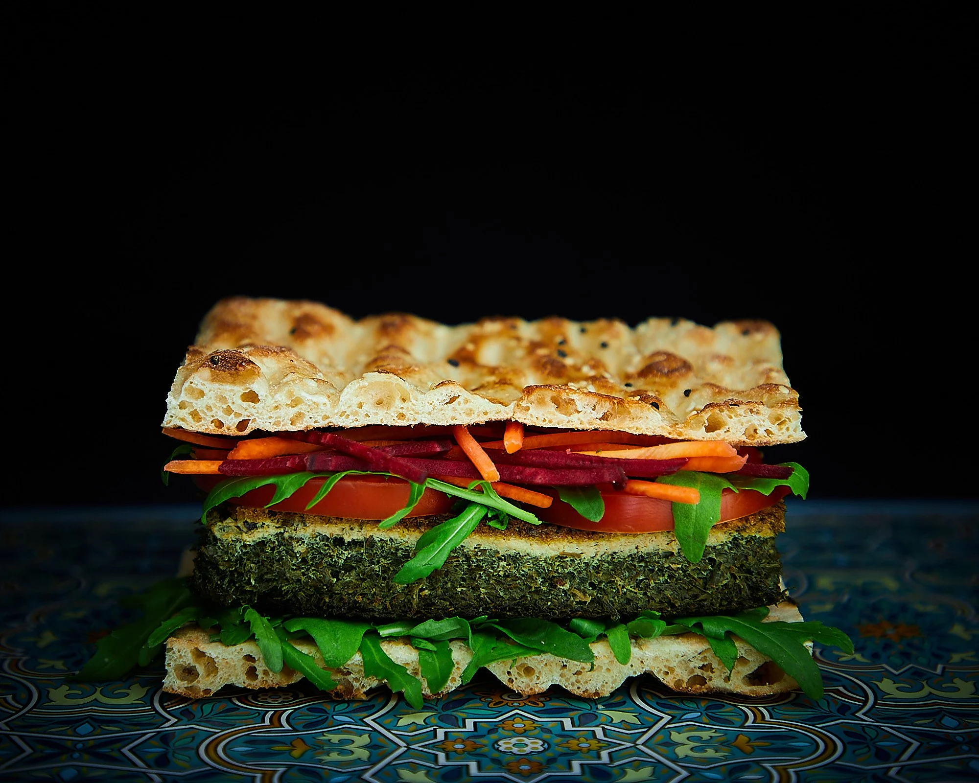 Vegetarian burger with dark mood lighting, containing arugula, tomato, shredded carrots, beet slices, veggie patty, sat on a patterned tile surface. Sandwich photography, dark and moody food photography. 