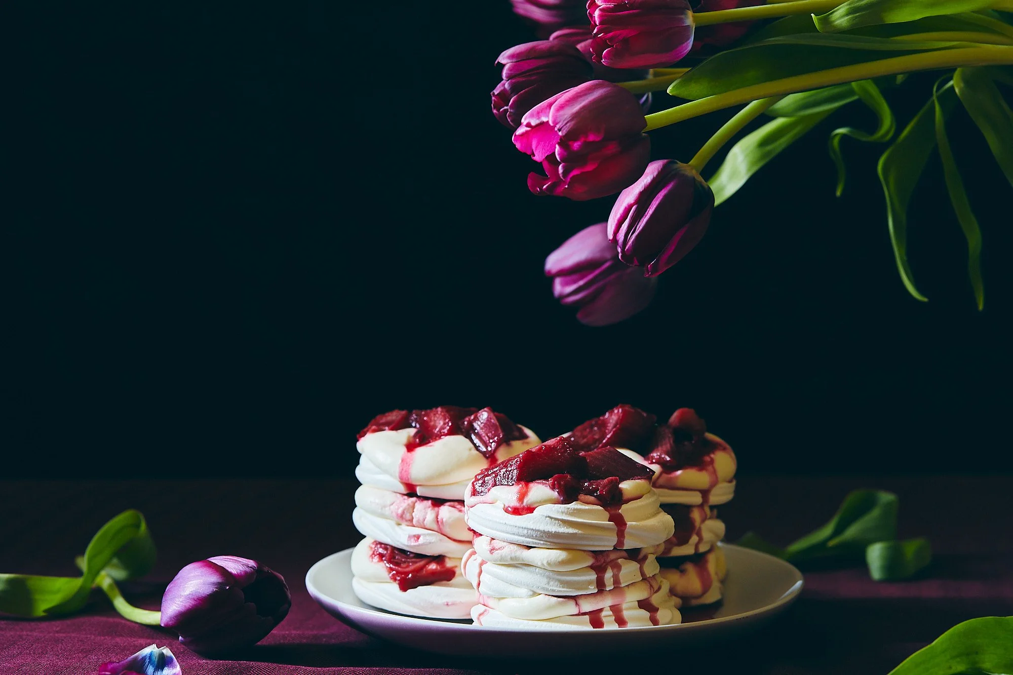 Stack of meringue cookies topped with berry compote on a white plate, with purple tulips hanging above and around on a dark background. Dessert photography, bakery photography, photo taken by Mah vafaei. 
