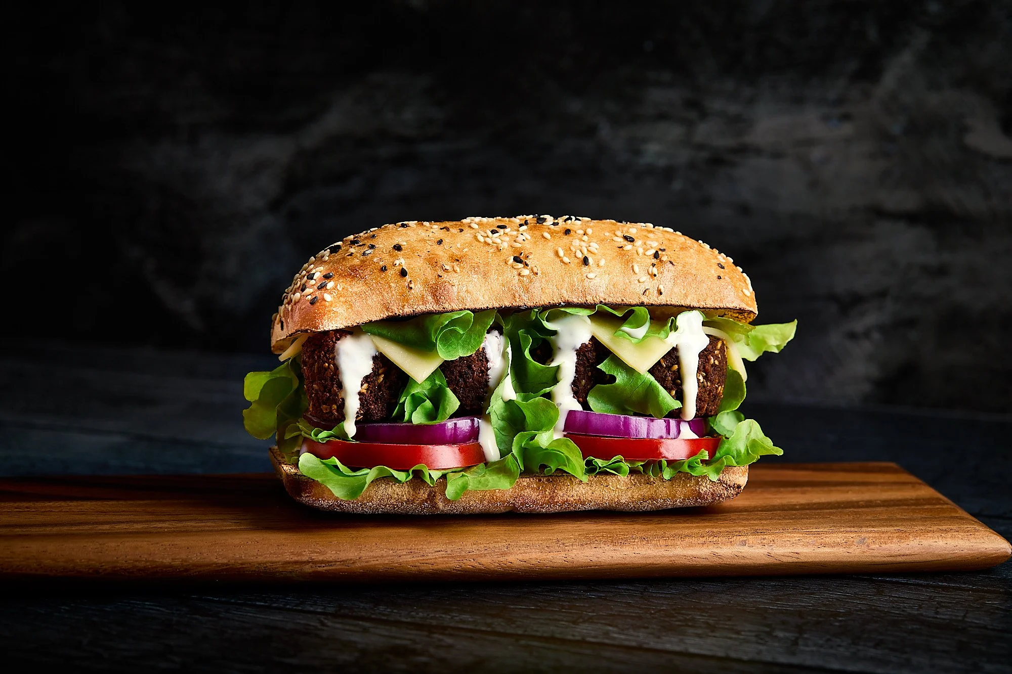 A vegetable burger with lettuce, tomato, onion, cheese, a veggie patty, and sauce on a sesame seed bun, placed on a wooden board against a dark background. Cafe photography, food photography, dark and moody food photo.