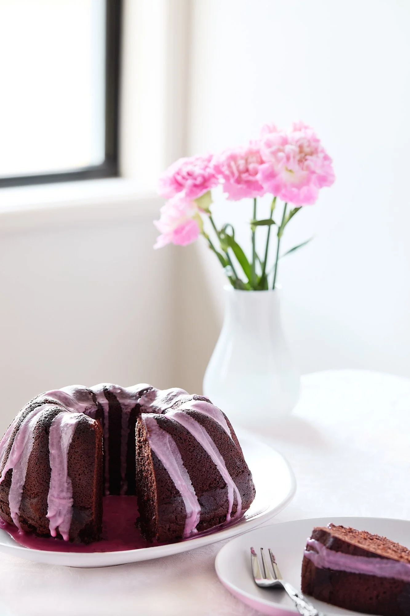 Chocolate bundt cake with pink icing on a white plate, a slice on a smaller plate with a fork, and pink carnations in a white vase on a white table near a window. Dessert photographer, bright food photograph. 