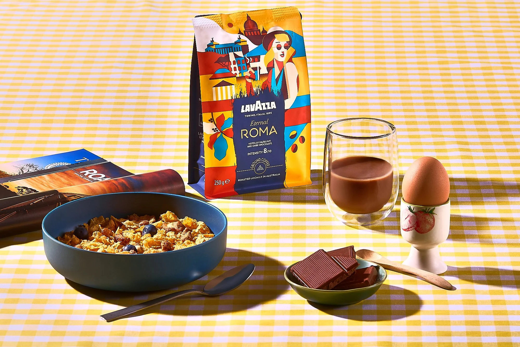 Breakfast table with cereal, chocolate, eggs, coffee, and a magazine on a yellow checkered tablecloth. Magazine photography, product photographer. Lavazza coffee brand photography.