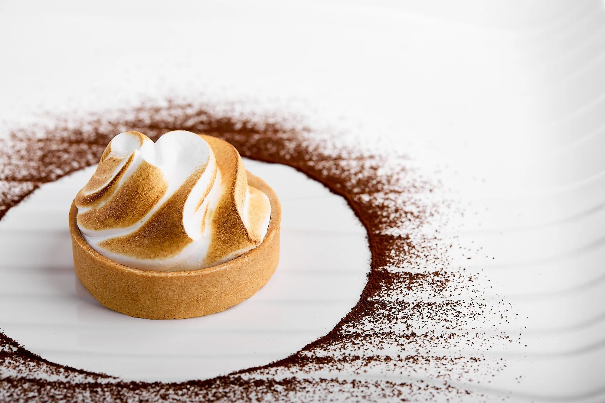 A tart with a baked meringue topping on a white plate, decorated with a circle of cocoa powder. Dessert photographer in Sydney, patisserie photography.  