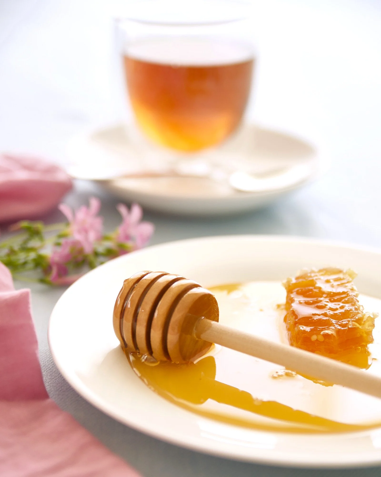 Close-up of a white plate with honey and a honey dipper, with a piece of honeycomb, a glass of tea, pink flowers, and pink napkin in the background. Table scape photo, food photography 
