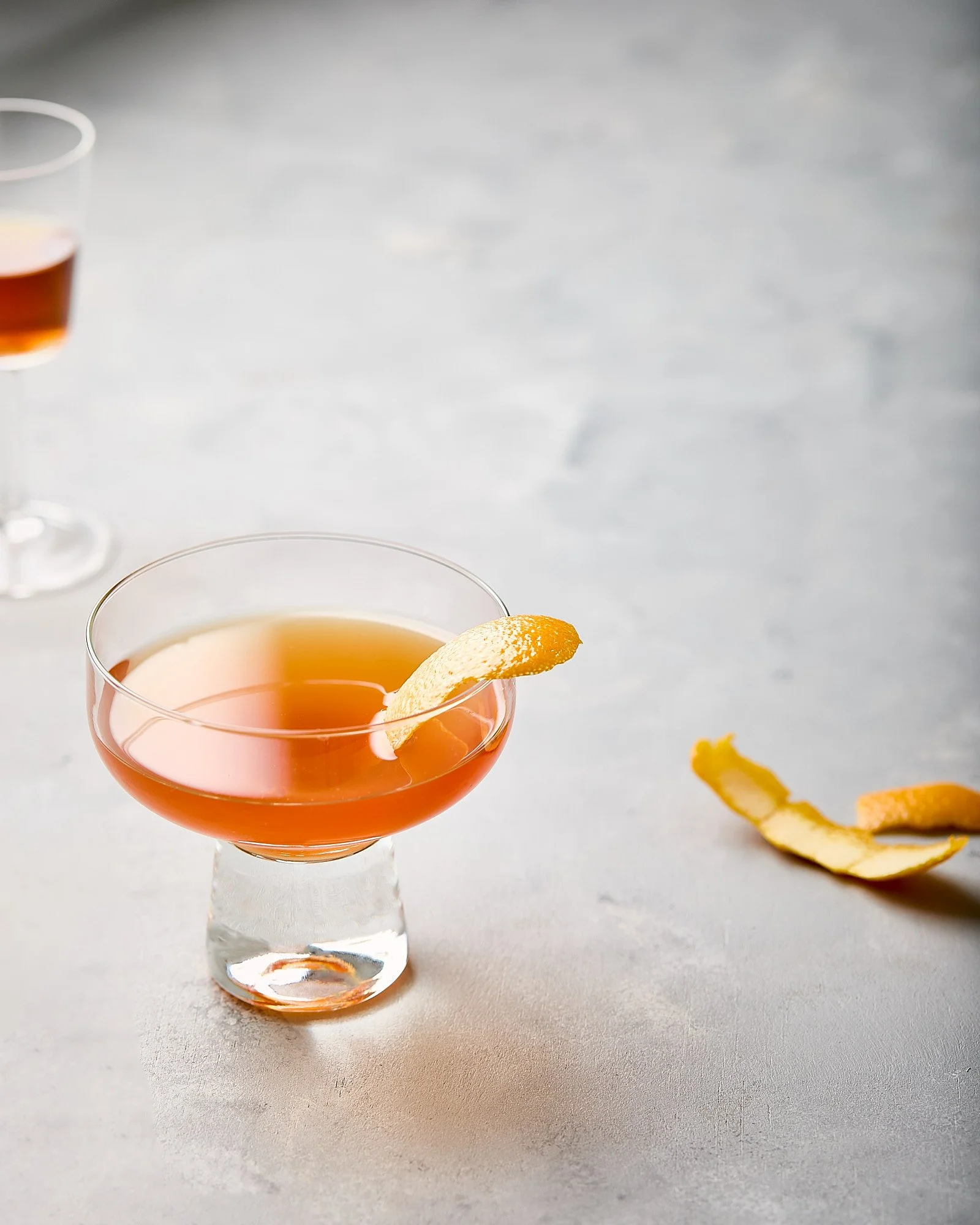 A cocktail glass with pinkish liquid garnished with a lemon peel, placed on a light surface. There is a second similar glass in the background and lemon peel strips on the right. Beverage photography, drink photographer, photo taken by Mah Vafaei.