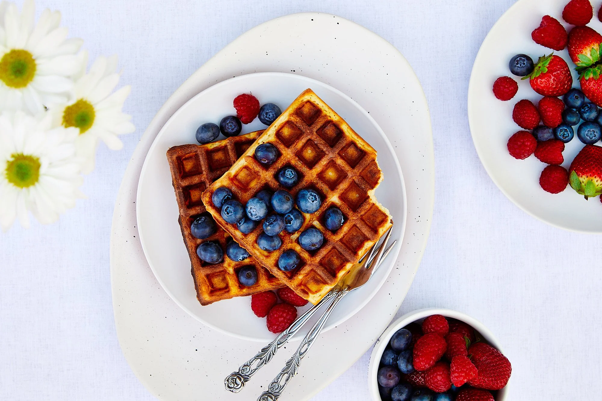 Two waffles topped with blueberries on a white plate, surrounded by bowls of mixed berries, with daisies nearby. Flatly food photography, cafe photographer, breakfast photo.