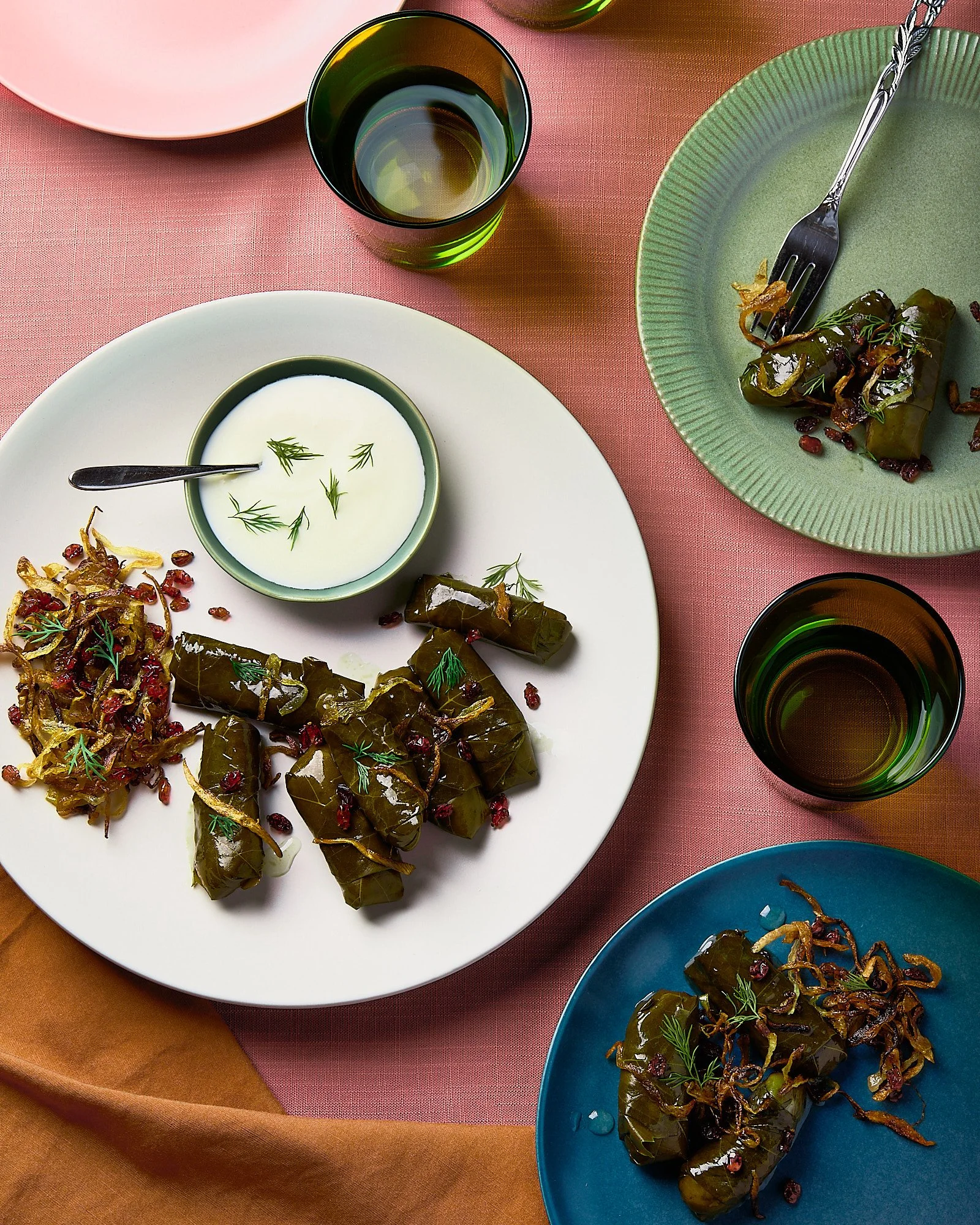 A top view of a table with plates of stuffed grape leaves, a bowl of yogurt with herbs, and glasses of water on a pink tablecloth. Flatlay food photography, restaurant photographer. 