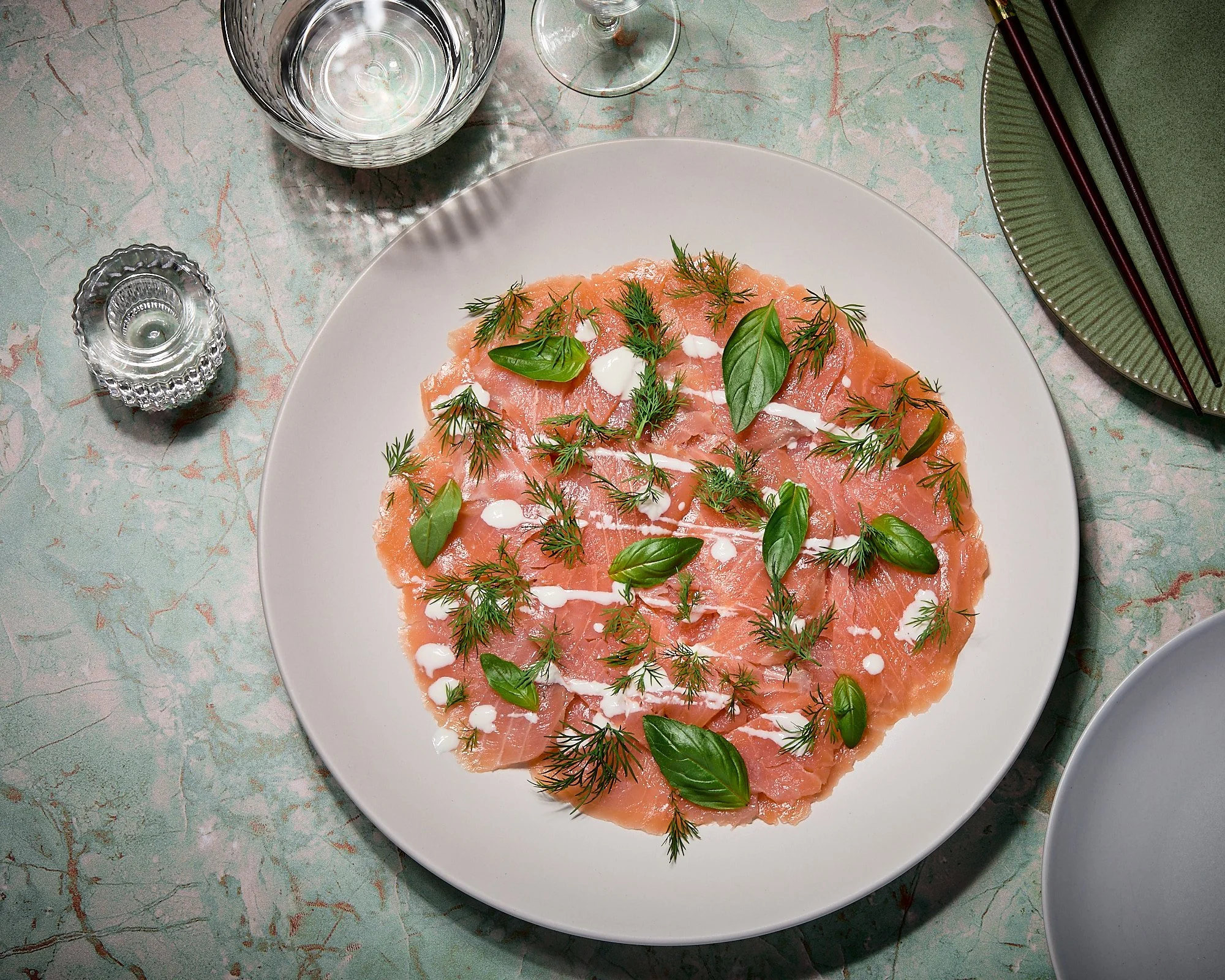 A plate of raw sliced salmon garnished with fresh herbs and drizzled with a white sauce, set on a table with glasses of water, a small candle holder, a green plate with chopsticks, and an empty bowl. Flatly photo, food photography