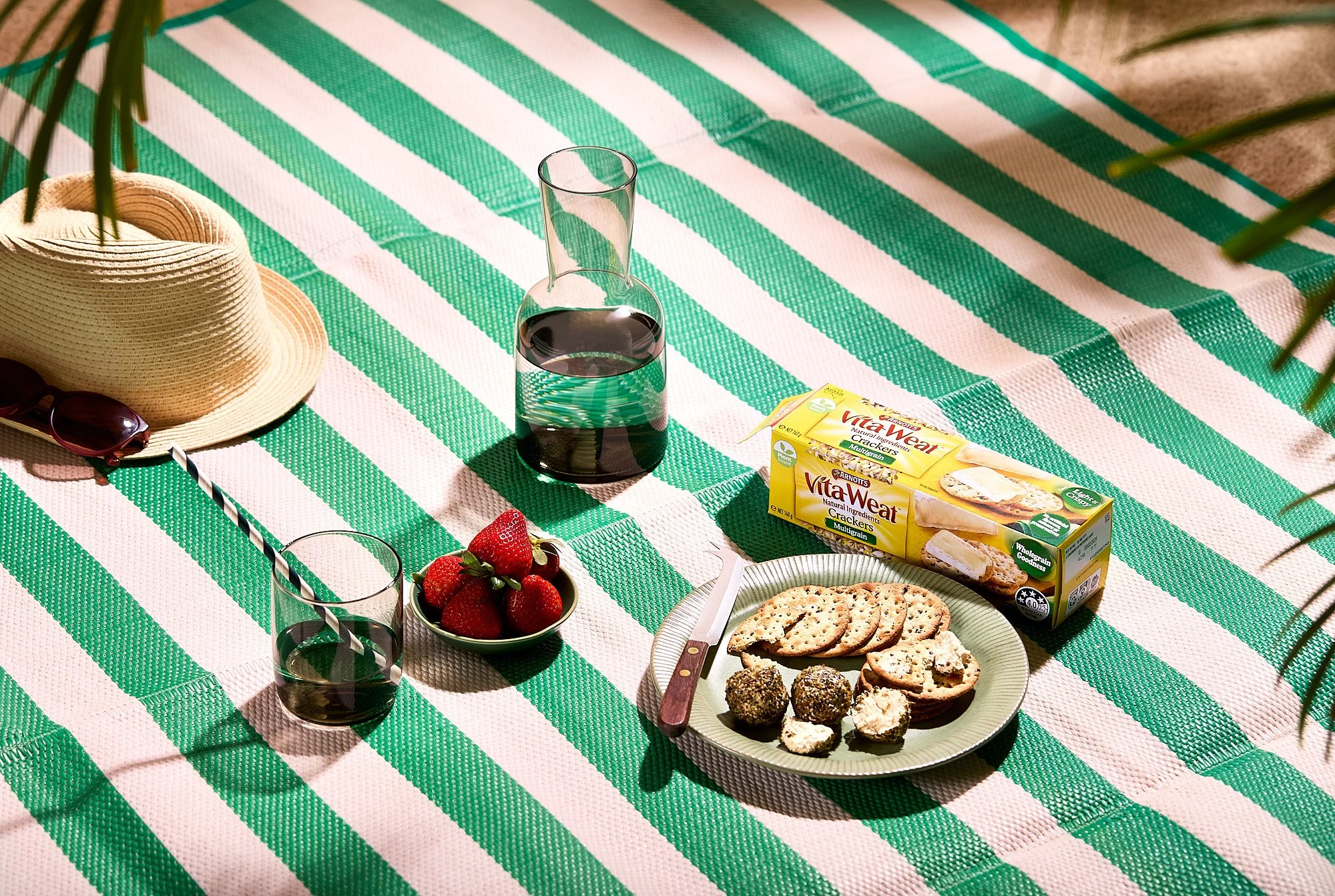 Picnic blanket with strawberries, a glass of dark soda with a striped straw, a box of VitaWeat crackers, a dish with assorted cookies and brownie bites, a straw hat, and sunglasses, with palm leaves at the edges. Commercial food photography. 