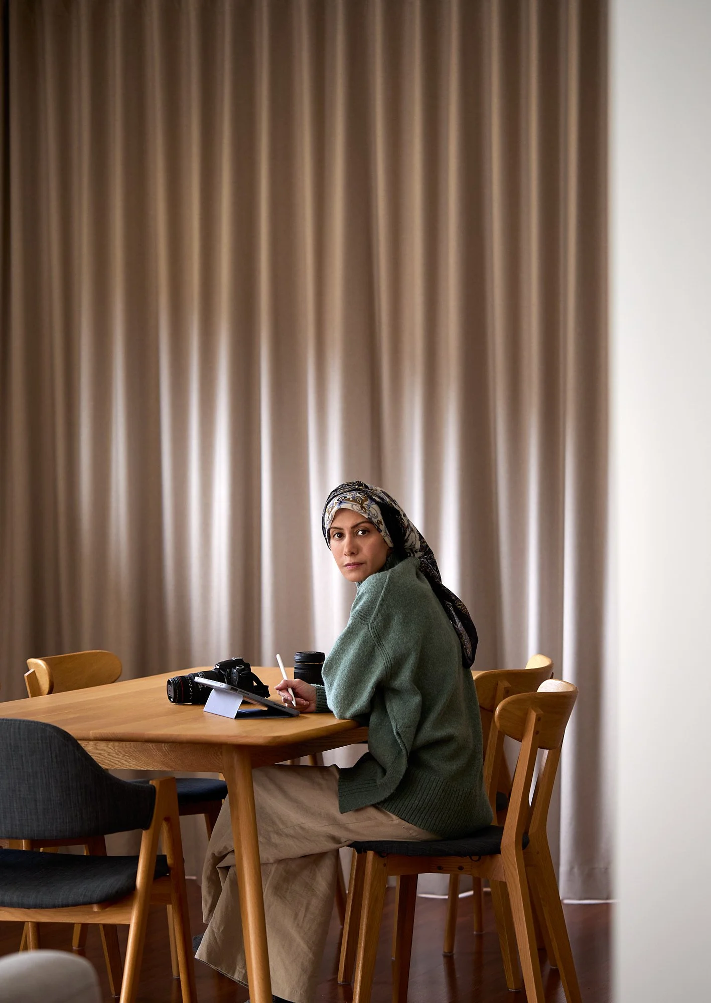 A woman sitting at a wooden dining table with arms resting on it, holding a stylus and looking at the camera, with camera and lenses in front of her, beige curtains in the background. Mah Vafaei, commercial brand photographer.
