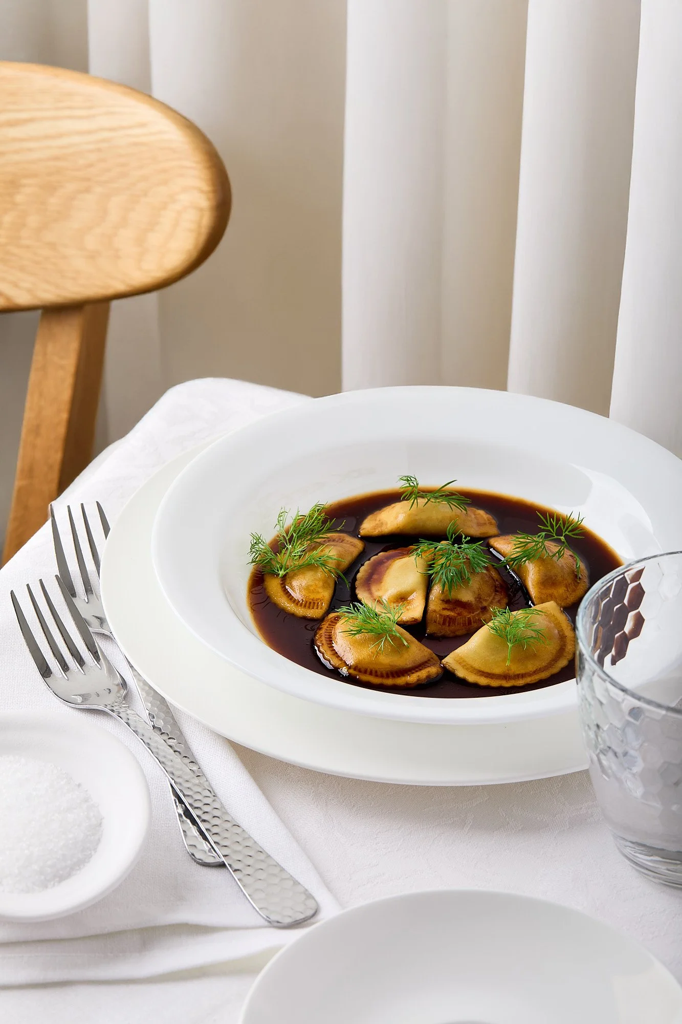 A white plate of ravioli in a dark sauce garnished with green herbs on a dining table. Luxury restaurant photography, brand photographer. 