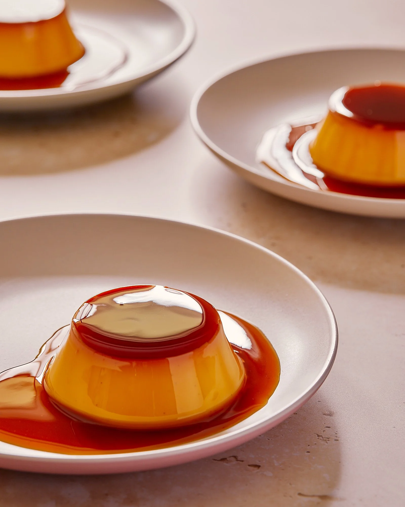 Three white plates with caramel flan desserts topped with caramel sauce on a light-colored surface. Dessert photography, patisserie photographer. 