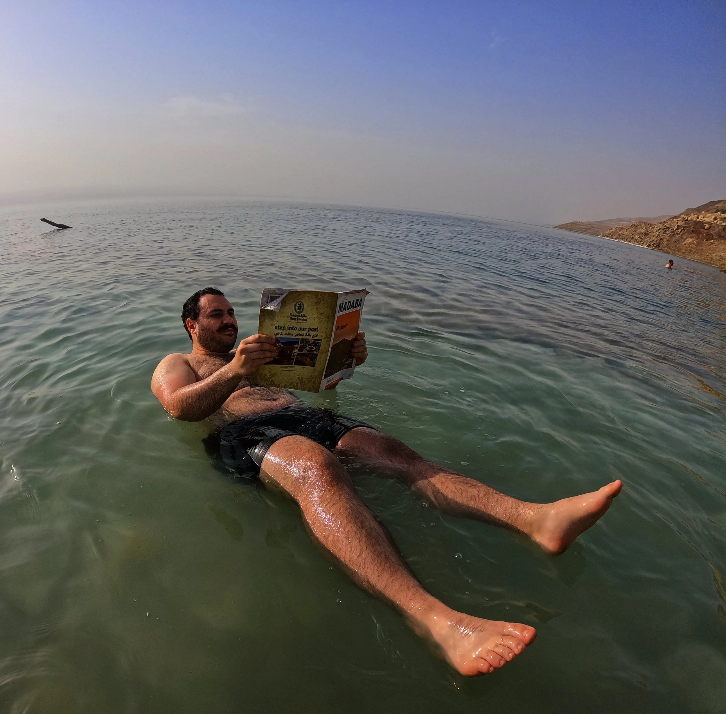 Homem relaxando na água do mar morto, deitado de costas, lendo um jornal, com o mar ao fundo e céu claro.