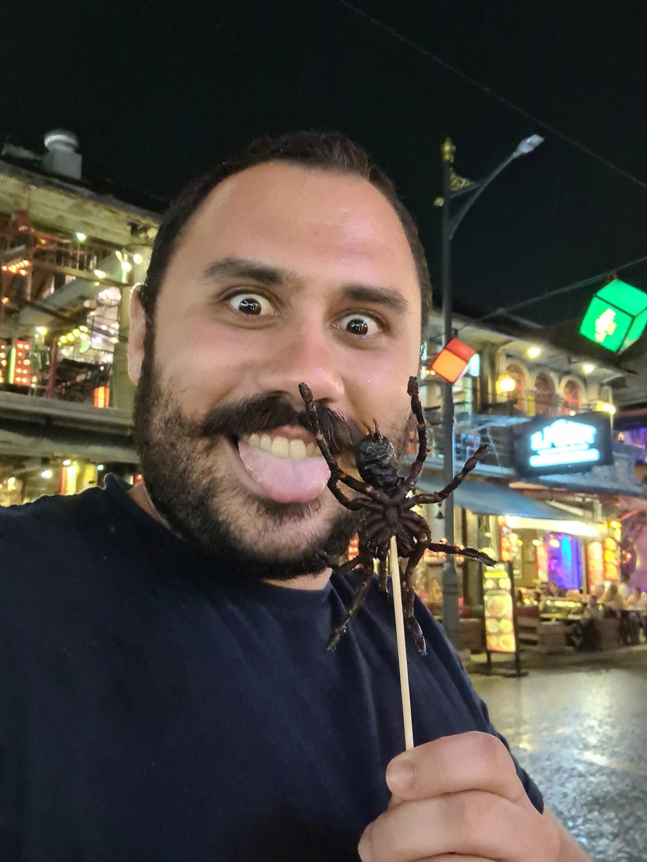 Homem prestes a comer uma espetada de aranha como snack nocturno na cidade de Siem Reap (Camboja), com luzes e lojas ao fundo.