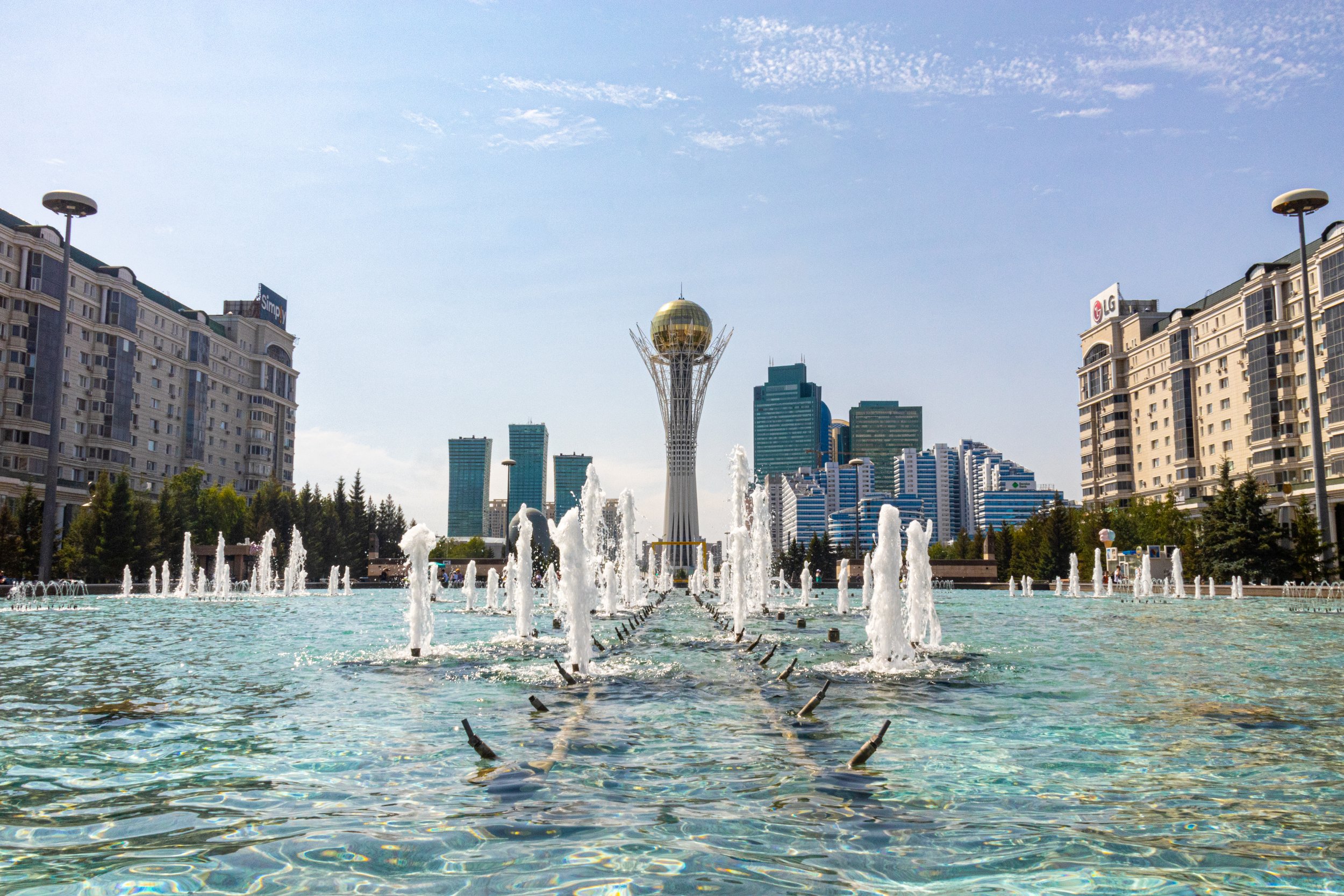 Vista da Torre Bayterek em Astana, Cazaquistão, com fontes e edifícios modernos ao redor, sob céu claro.