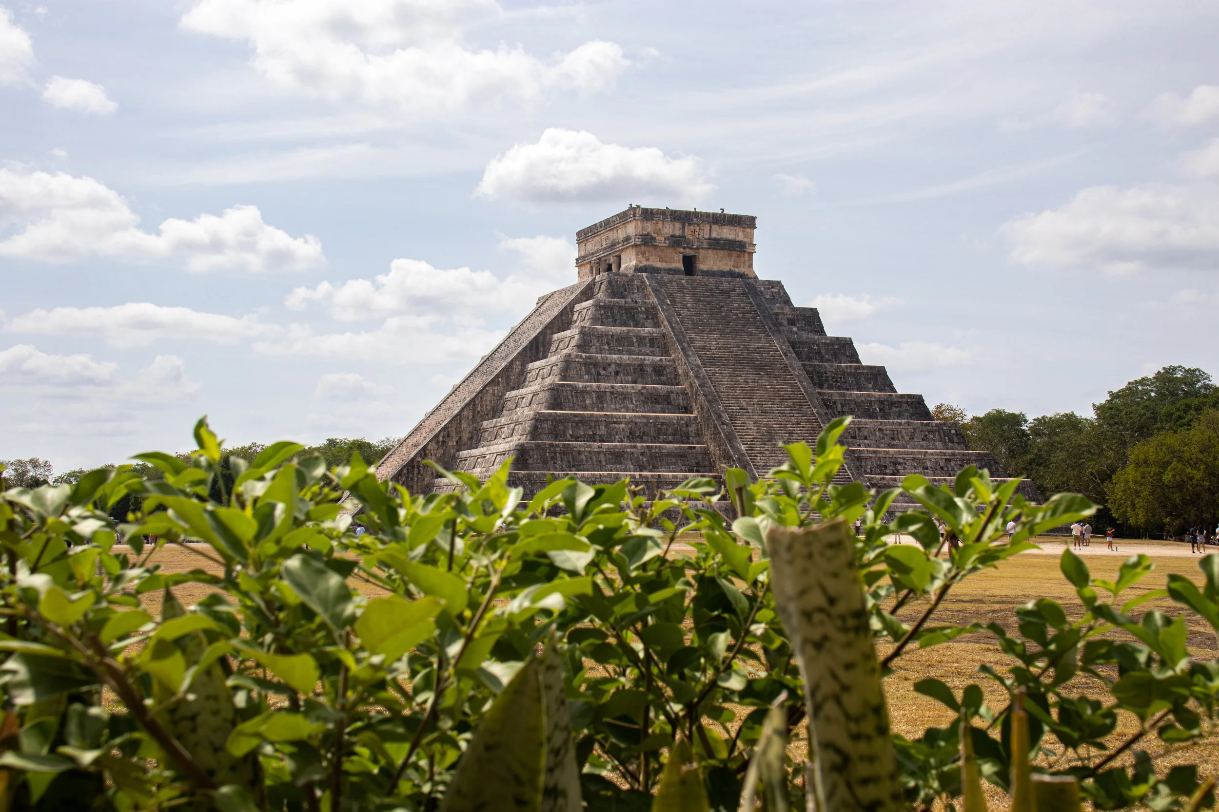 Pirâmide de Chichen Itza com vegetação no foreground e céu com nuvens ao fundo.