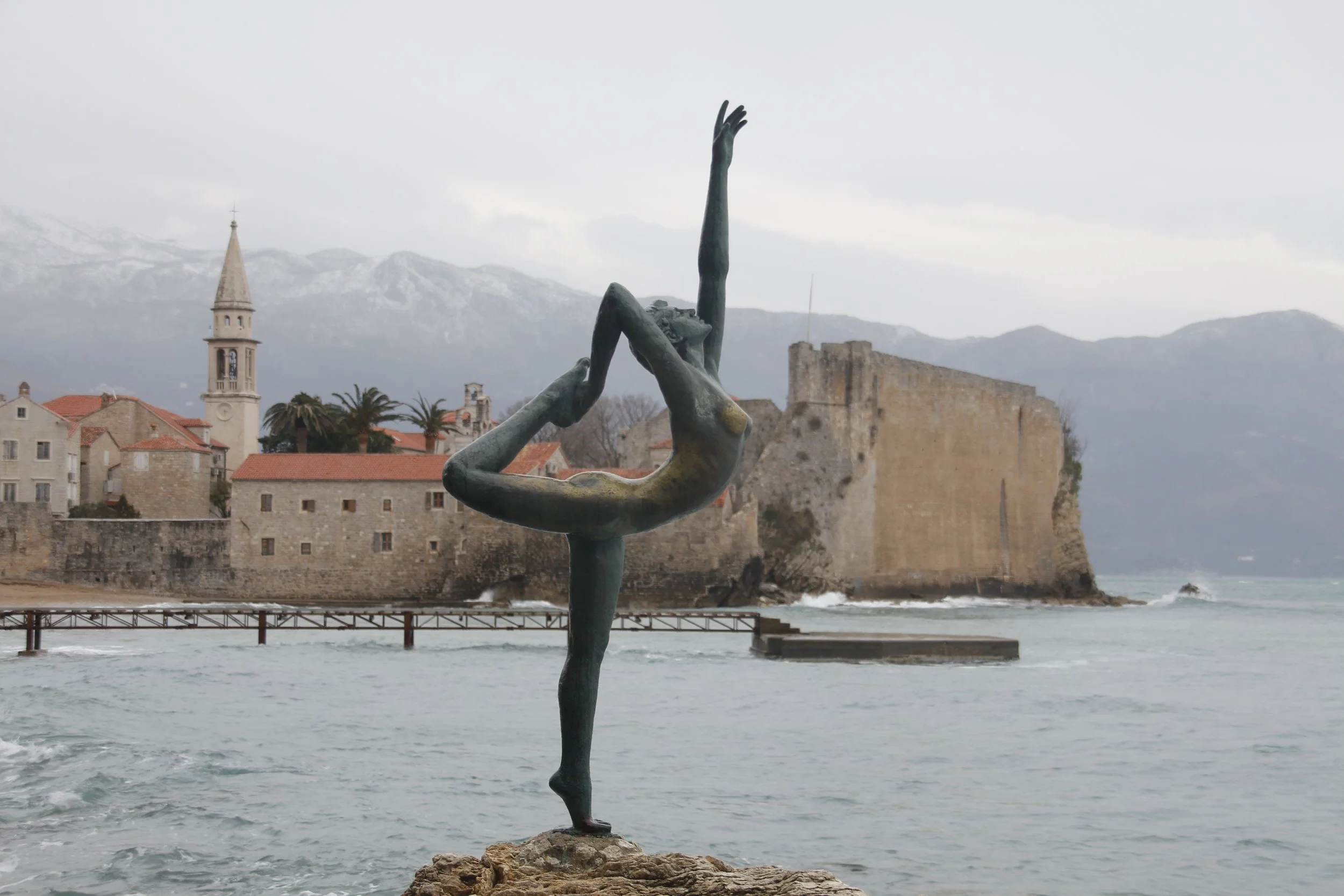 Escultura de uma bailarina de bronze na orla de uma cidade costeira (Budva, Montenegro), com muralhas antigas e montanhas ao fundo, em dia nublado.