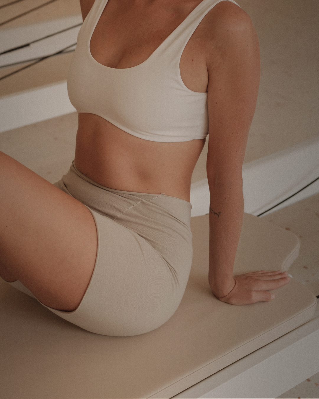 A woman sitting on a bench, wearing a white sports bra and beige shorts, with her one hand resting on the bench.