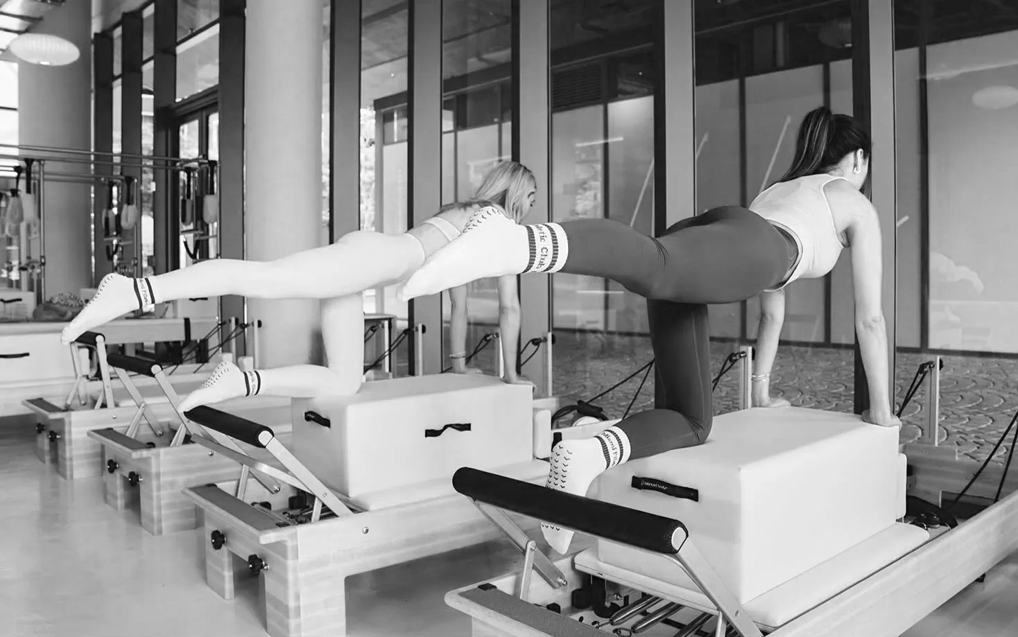 Two women practicing Pilates, using reformer machines in a modern fitness studio with glass walls.