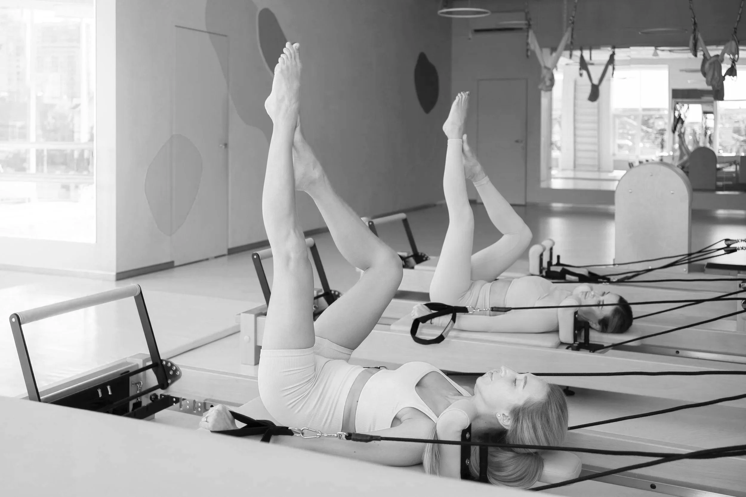 Two women lying on Pilates reformer machines, exercising with their legs raised and holding straps, in a gym studio.