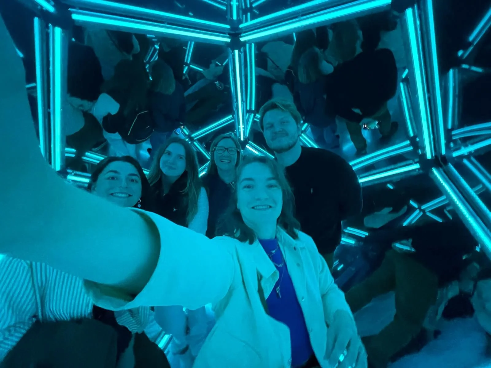 Group of five people taking a selfie inside a mirrored, illuminated tunnel with blue neon lights.