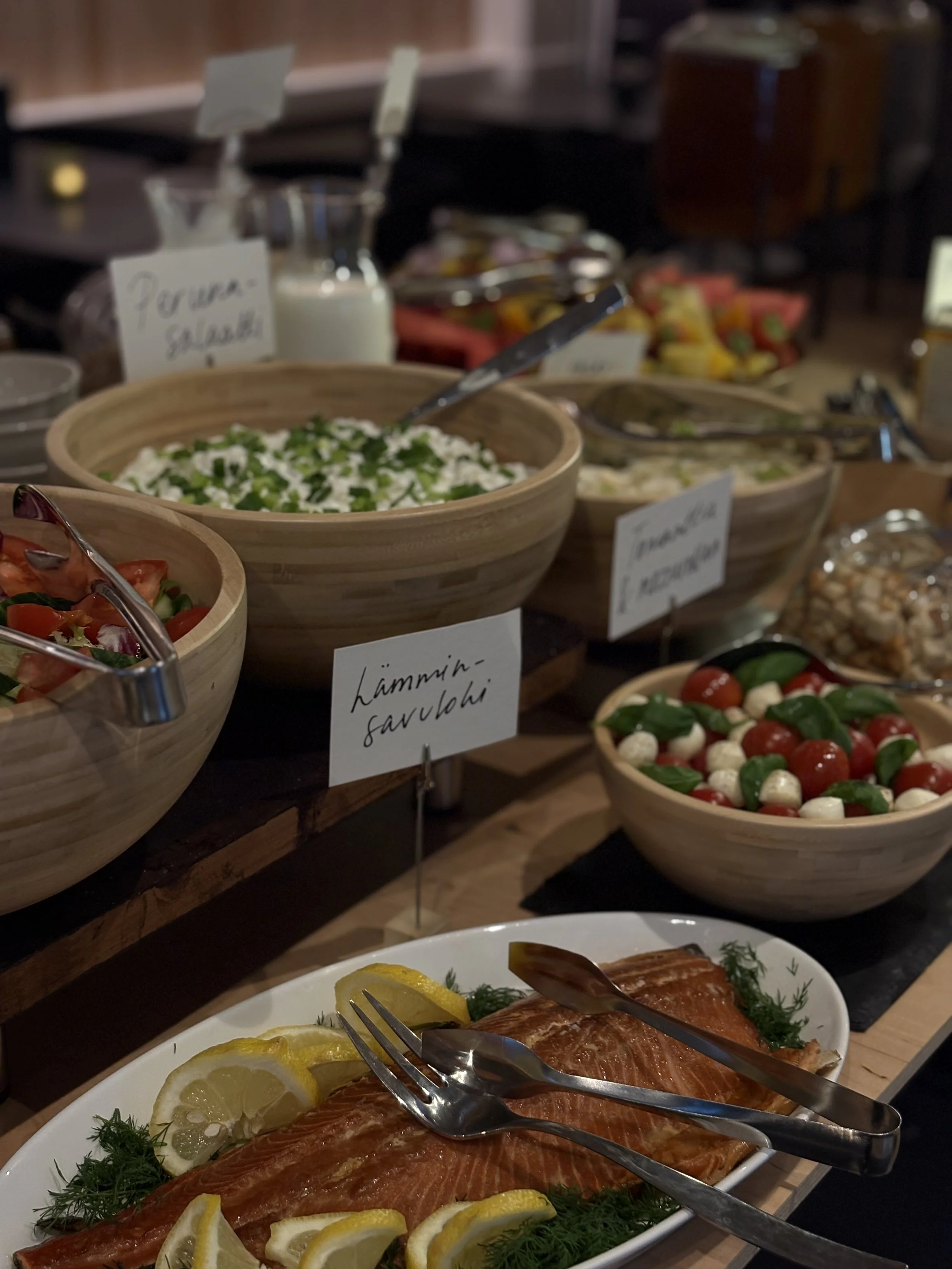 JAM Café & Bar, Nokia brunssipöydän kuva jossa on erilaisia salaattibowl-tyyppejä, joissa on vihanneksia ja mozzarellapalloja, sekä lasikannussa salaatinkastiketta ja lautasella tarjolla lohta ja sitruunoita. Tekstissä lukee 'Lämminsavulohi'.