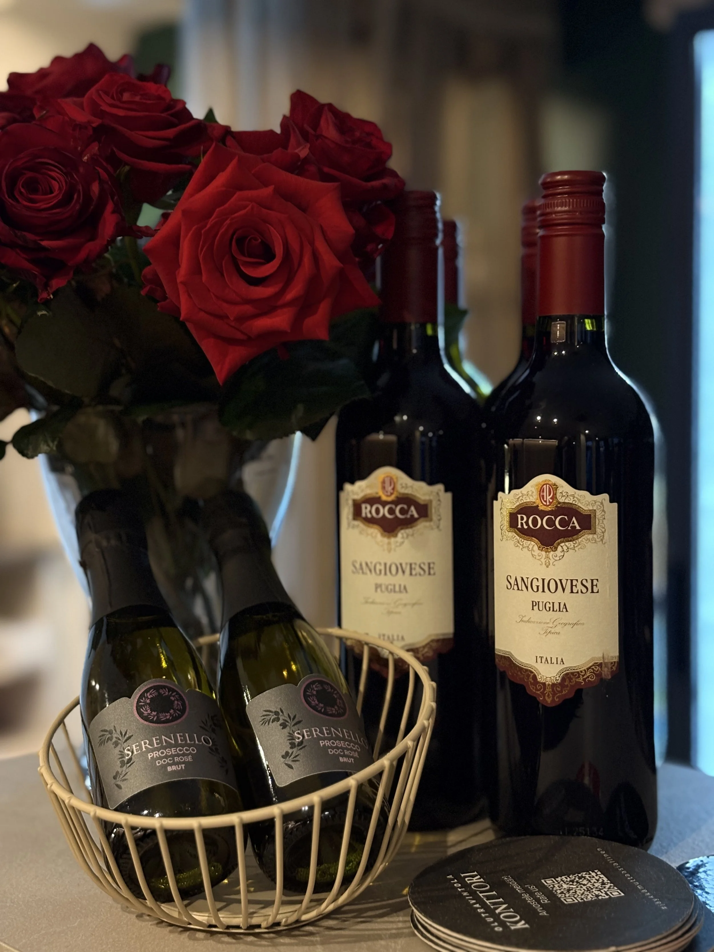 Vasemmalla punaisia ruusuja, keskellä kaksi pulloa punaviiniä ja oikealla kaksi pulloa roccia-sangiovese punaviiniä, alhaalla kori, jossa kaksi kivennäisvettä