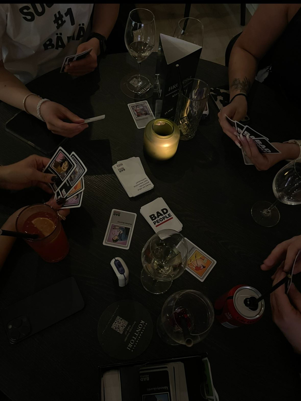 Ryhmä ihmisiä pelaa korttipelejä pöydällä, jossa on erilaisia juomia ja tarvikkeita, tunnelmallisessa kynttilän valossa.