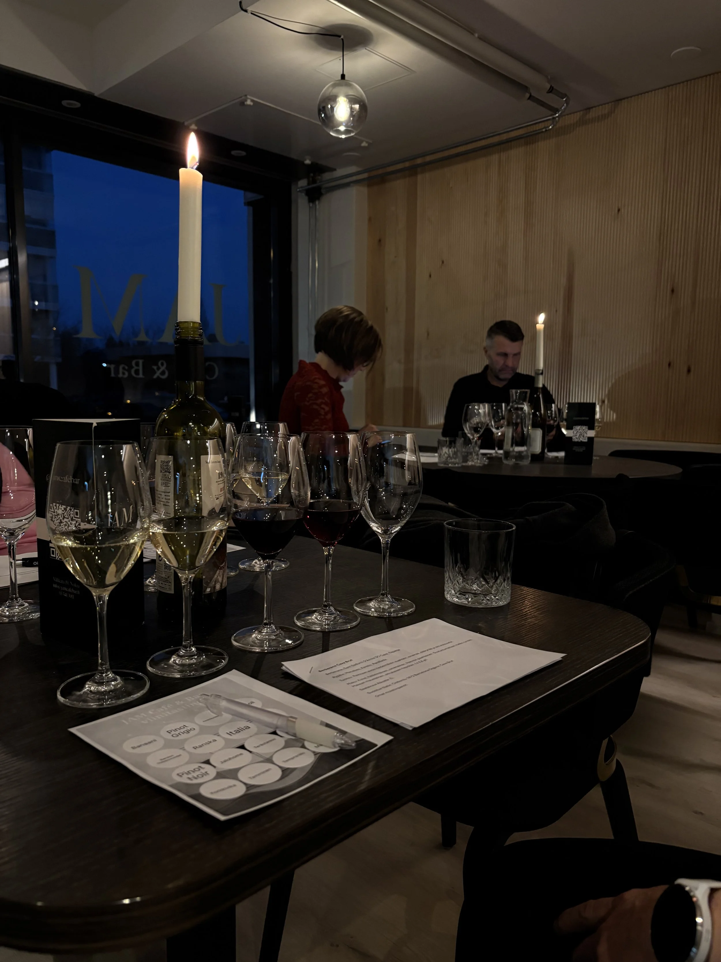 Ravintolassa pöytä, jossa viinilaseja ja viinibingo, kynttilöitä ja kaksi ihmistä taustalla, yksi nainen ja yksi mies. Valaistus on hämärää.