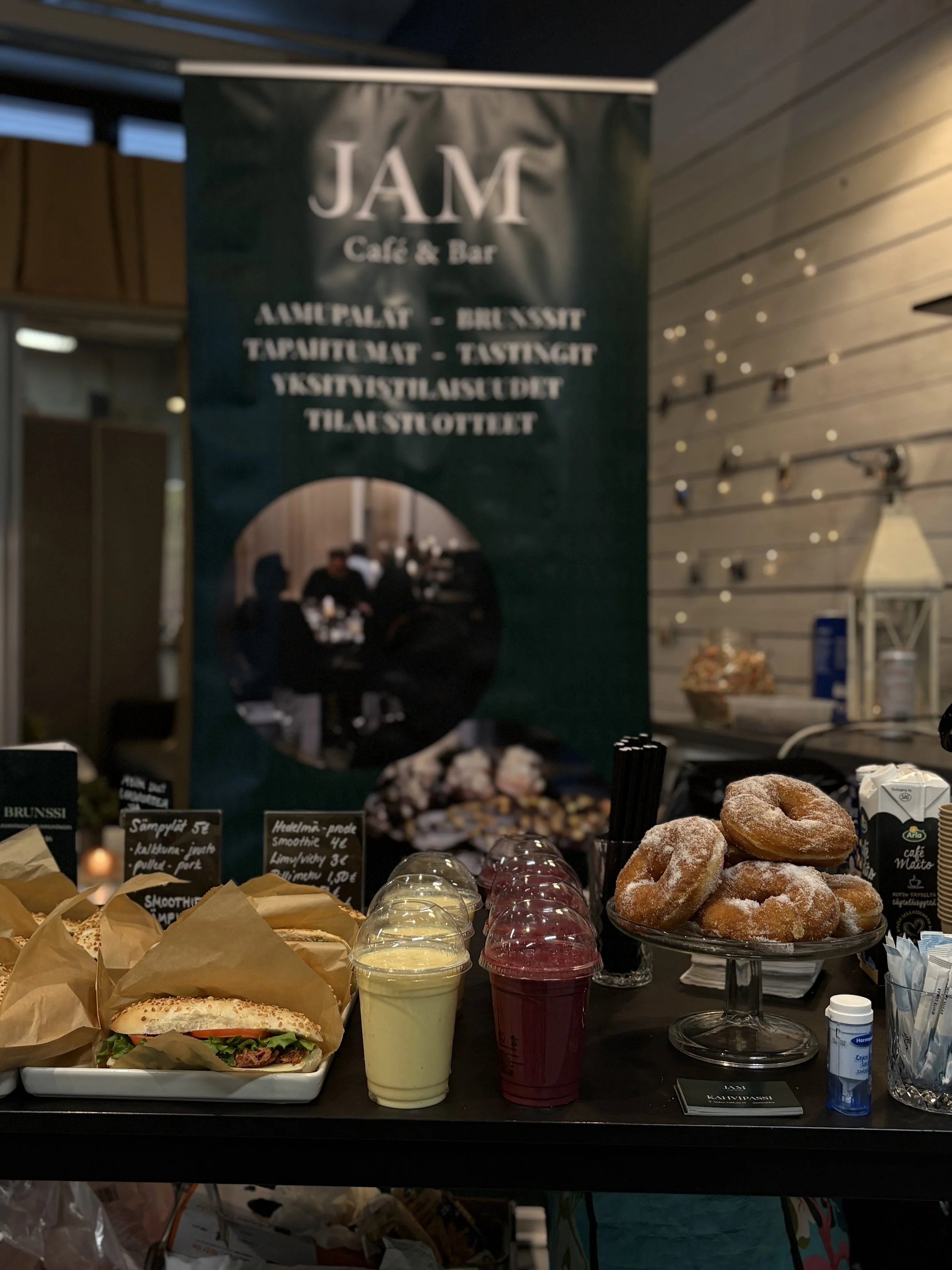 Messumyyntipiste, jossa on munkkeja, smoothiesja ja voileipiä myynnissä, vastapäätä suuri banneri ja tiski.