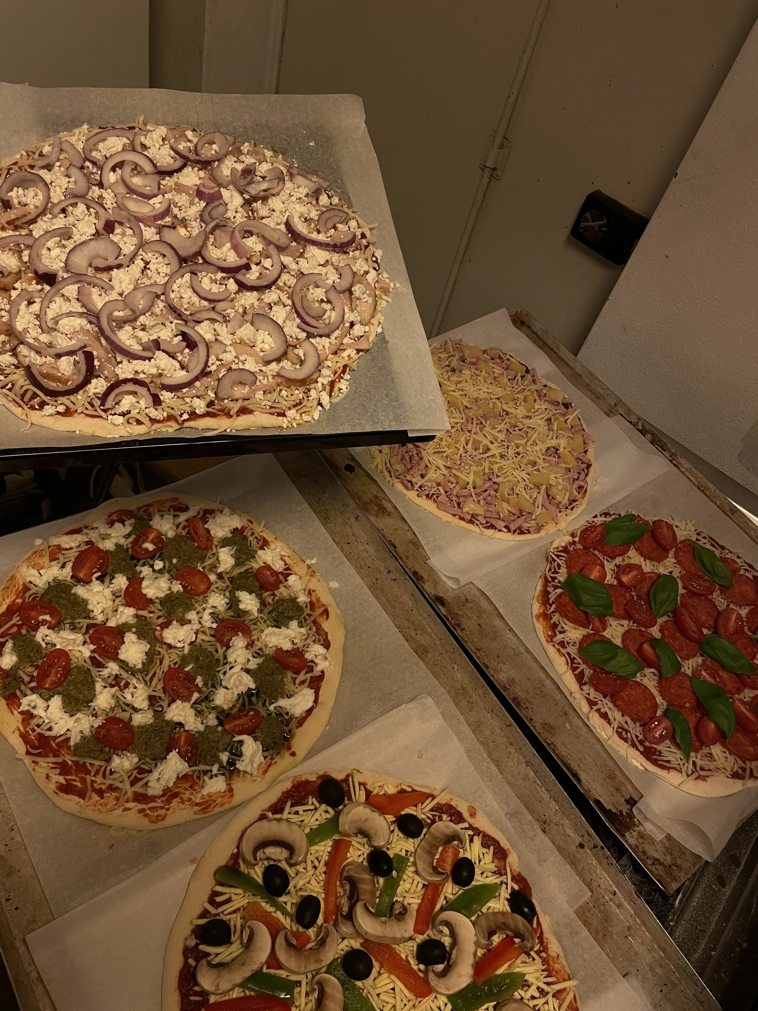 Useita erilaisia pizzatyyppejä uunissa, sisältäen kasviksia, juustoa ja lihaa, valmisteluvaiheessa pizza-teemapäivänä.