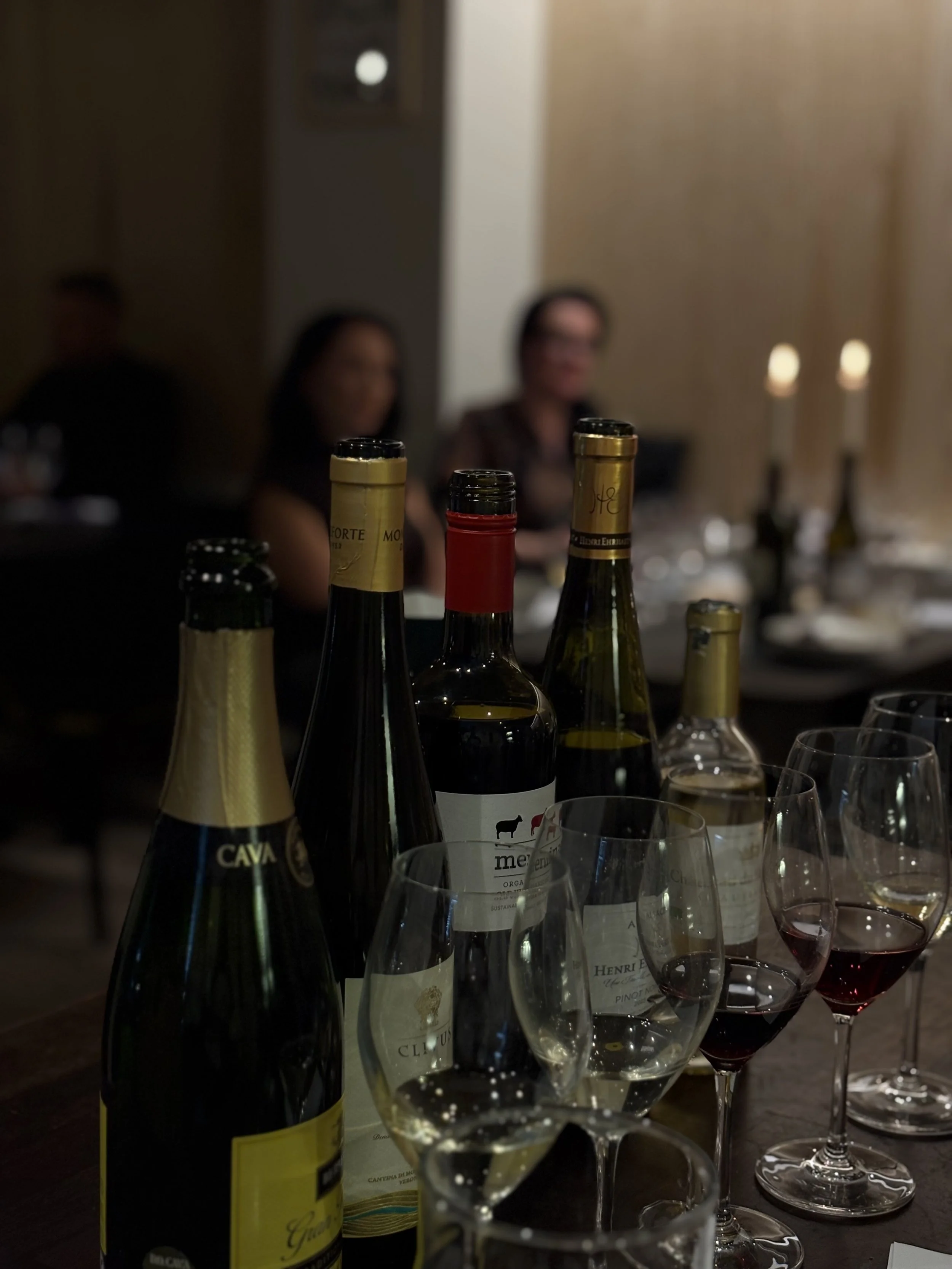 Kuva viinipulloista ja laseista pöydällä, teemana viinitilaisuus tai illallinen, taustalla häärii kolme ihmistä, mutta heidät on epätarkkoja.