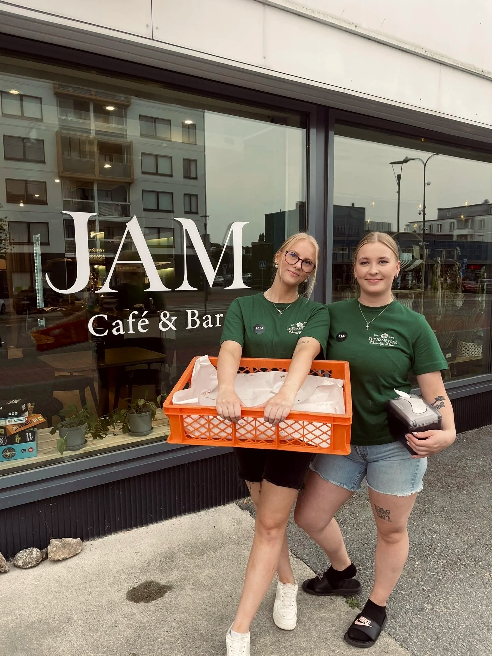 Kaksi nuorta naista seisovat ulkona JAM café & bar ravintolan ikkunan edessä, molemmat hymyilevät ja toisella on kori, toinen pitää kassia. He ovat pukeutunut vihreisiin T-paitoihin.