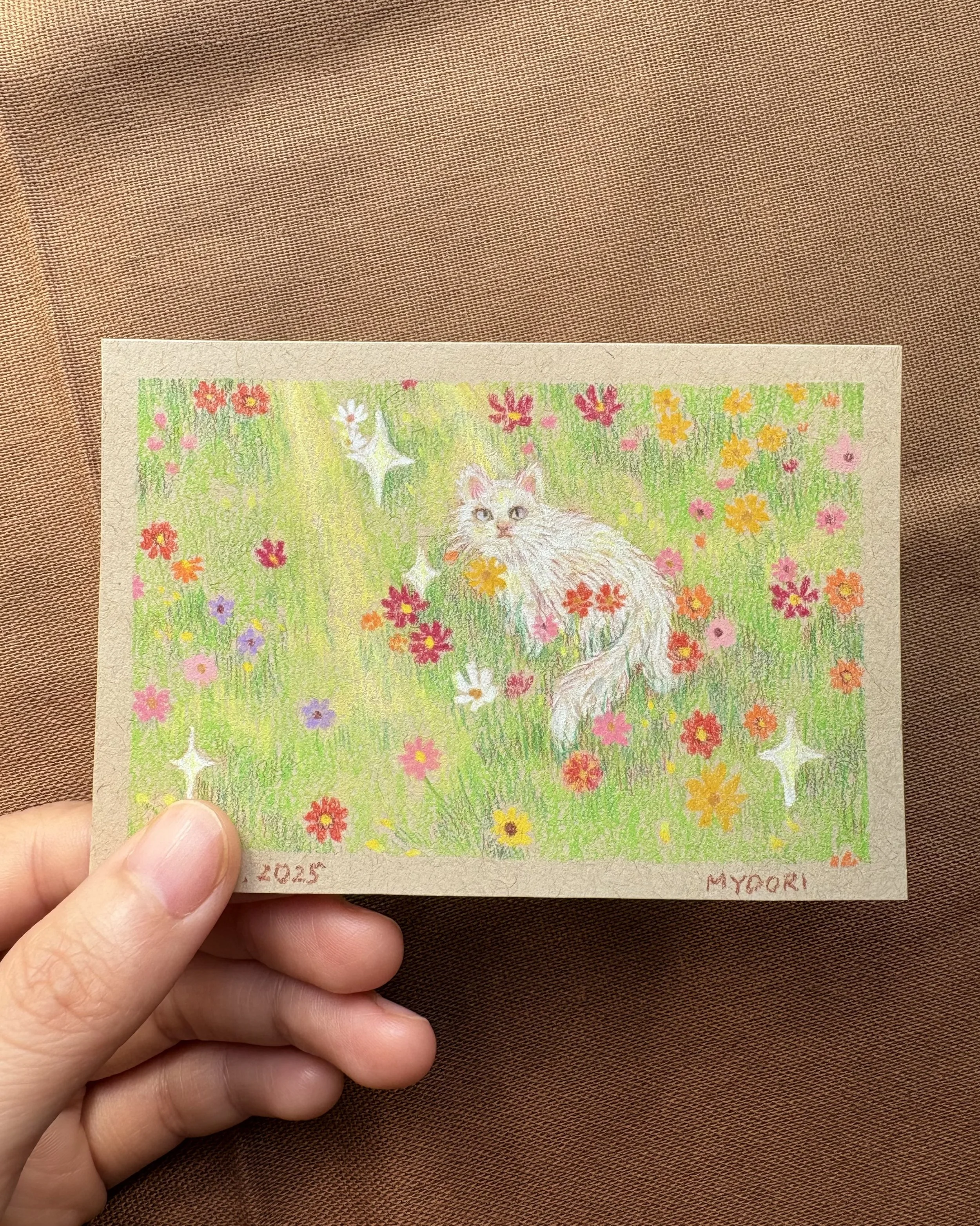 Colorful drawing of a white fluffy cat sitting in a field of pink, orange, purple, and white flowers with green grass and trees. Sparkles surround the scene. The bottom left corner has the date 2025, and the bottom right corner has the name MYOORI.