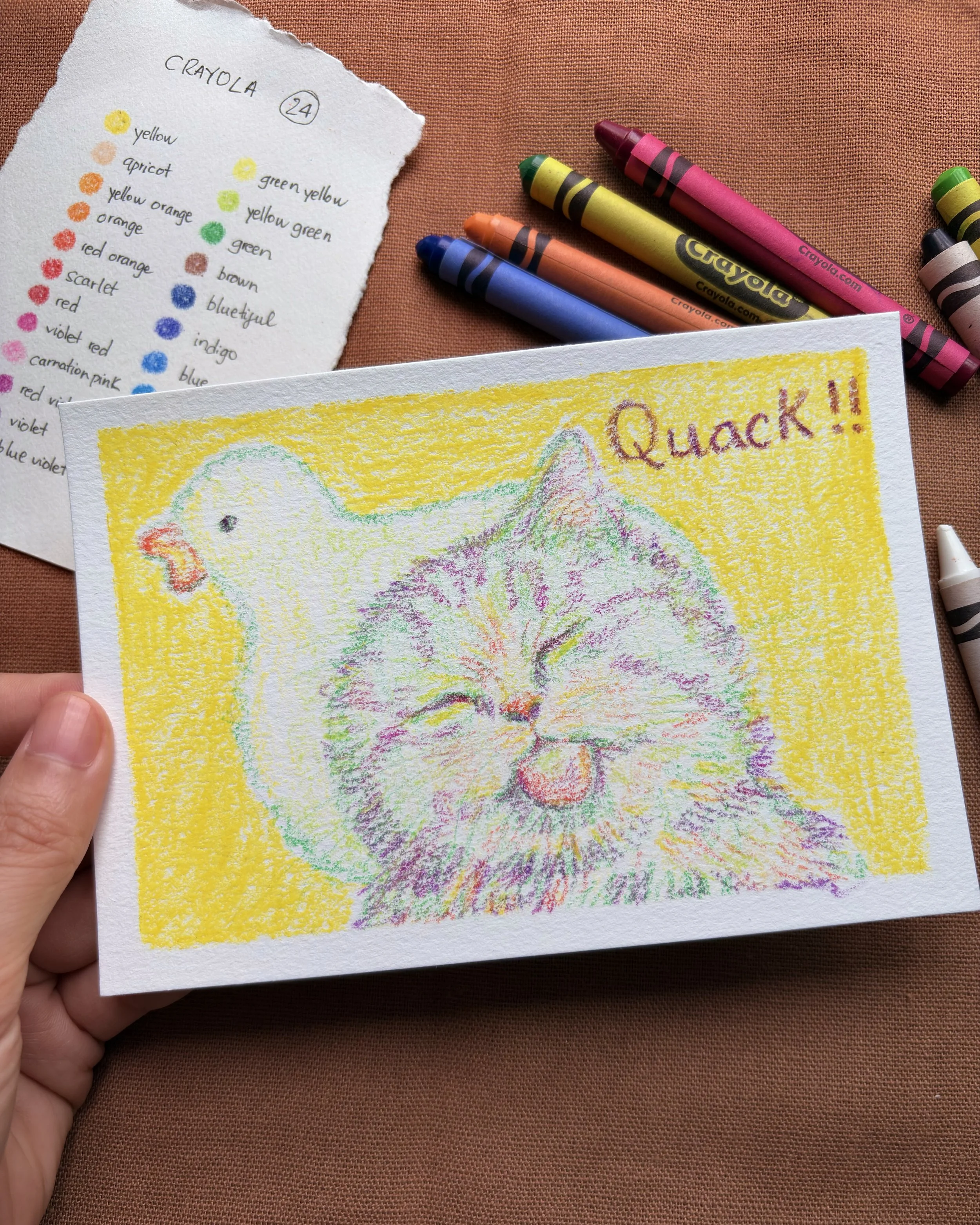 Colorful crayon drawing of a laughing cat with a chicken head behind it and the word "Quack!!" written in the top right corner, yellow background. Hand holding the drawing, with crayons and a color chart in the background.