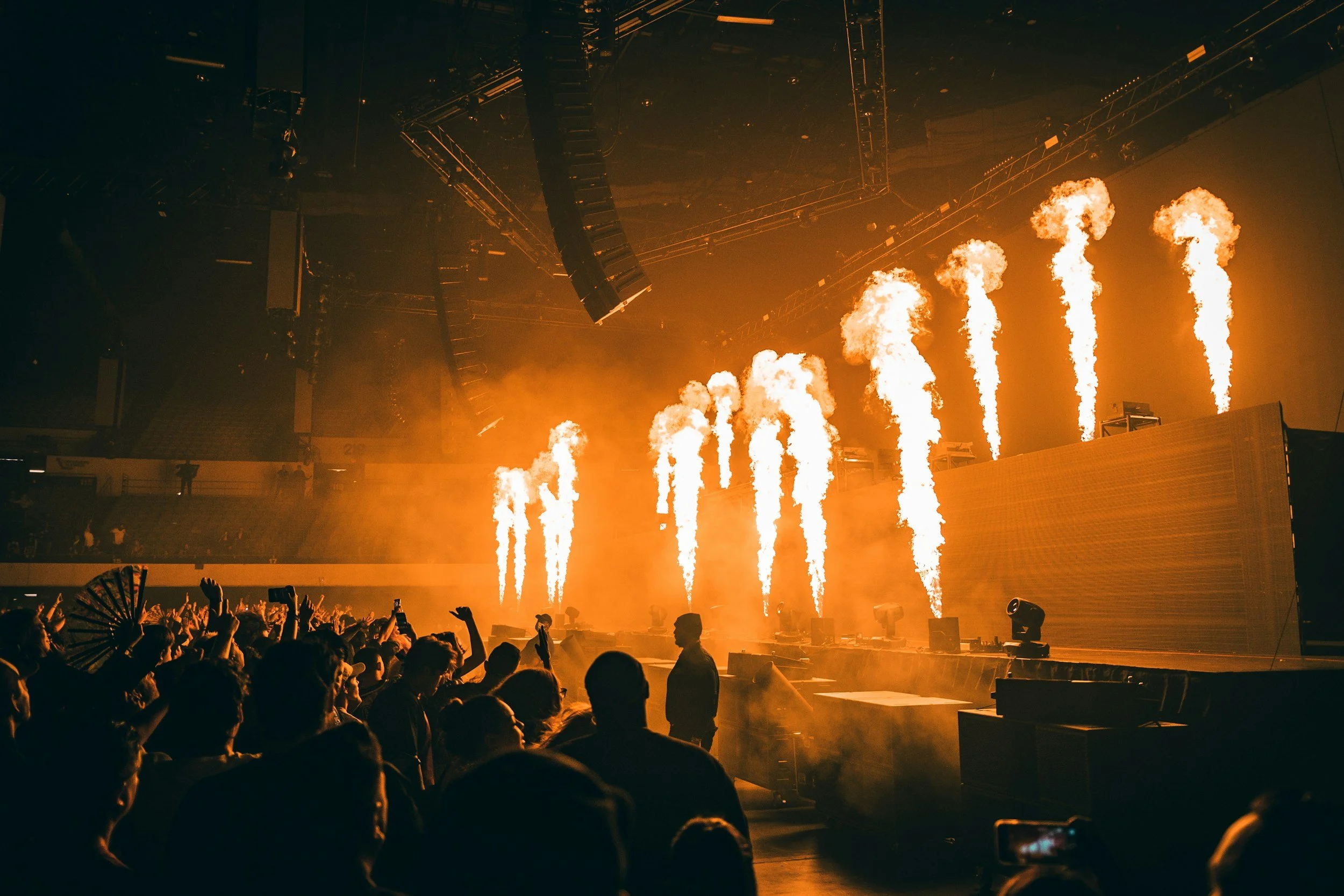 Crowd watching a concert with fire effects on stage, bright flames shooting up, and people raising their hands in excitement.