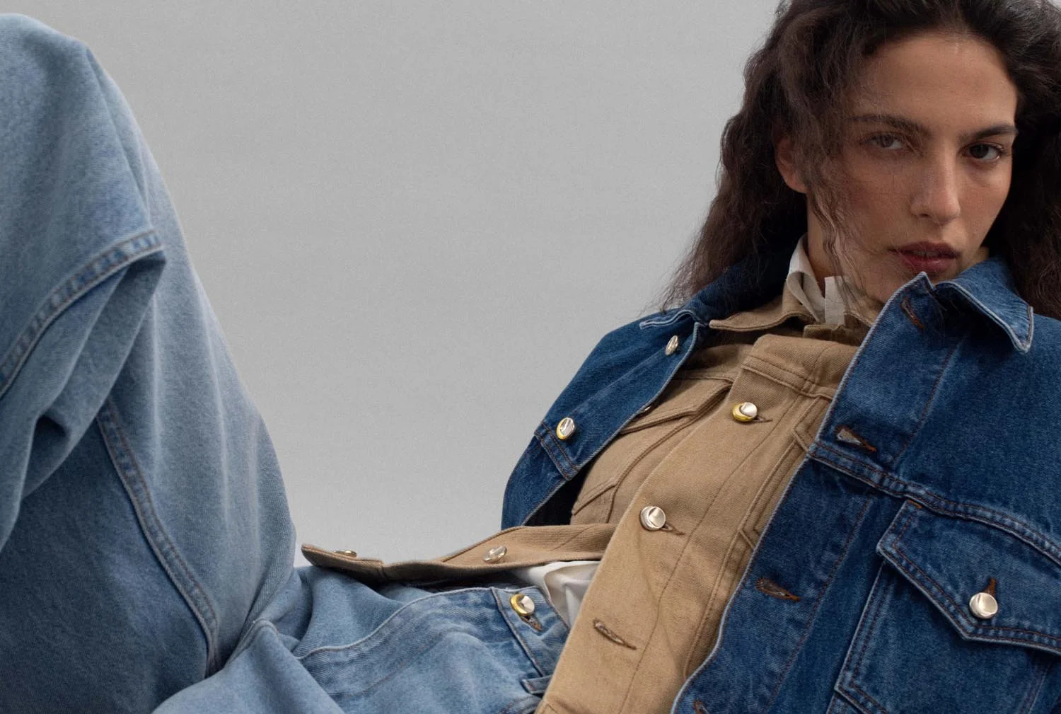 Nahaufnahme eines jungen Mannes in lässiger Kleidung, die einen schwarzen Parka und Jeans trägt, mit langem, lockigem Haar, der auf einem grauen Hintergrund sitzt.