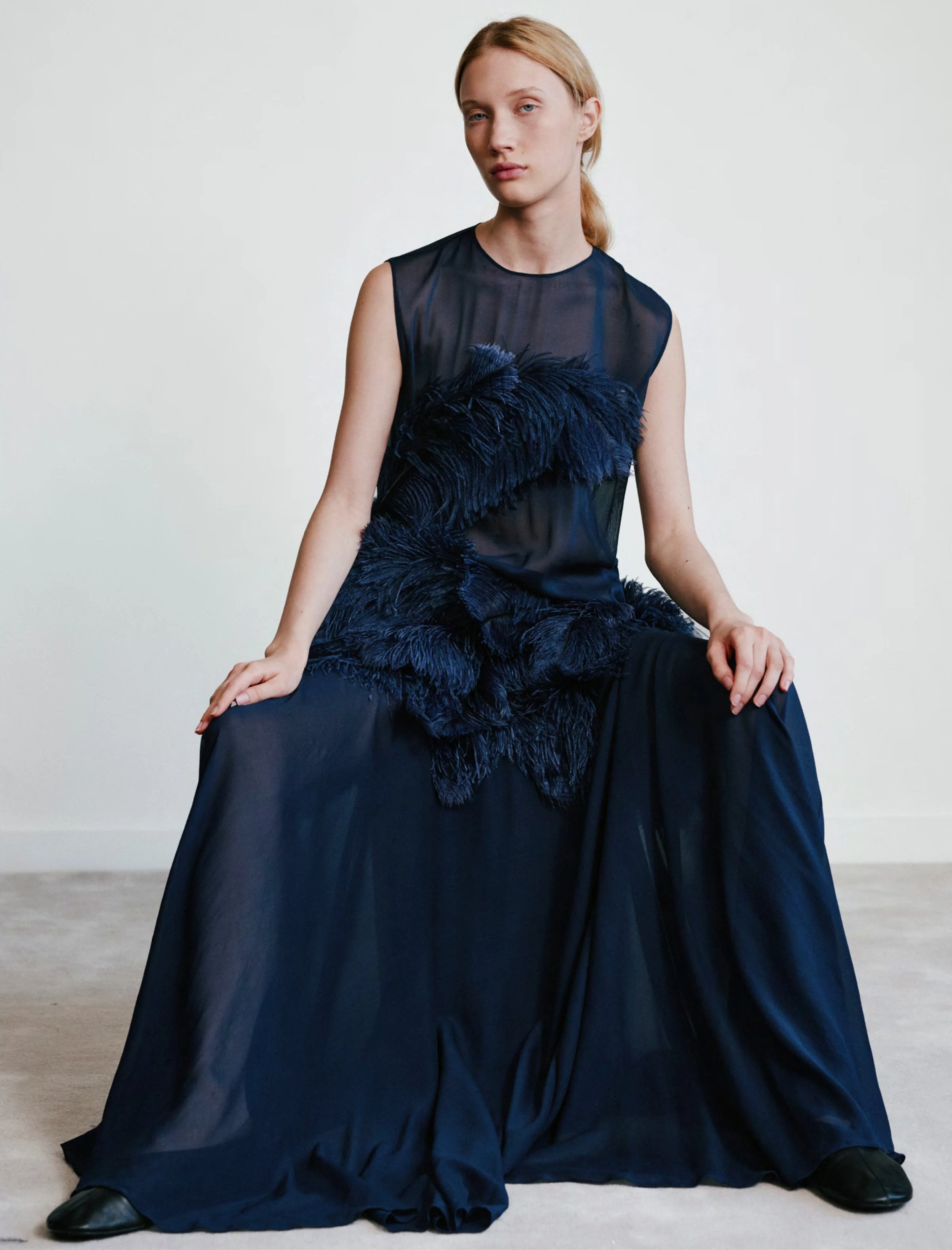 Frau in dunklem, eleganten Kleid mit Federn sitzend auf dem Boden vor weißem Hintergrund.