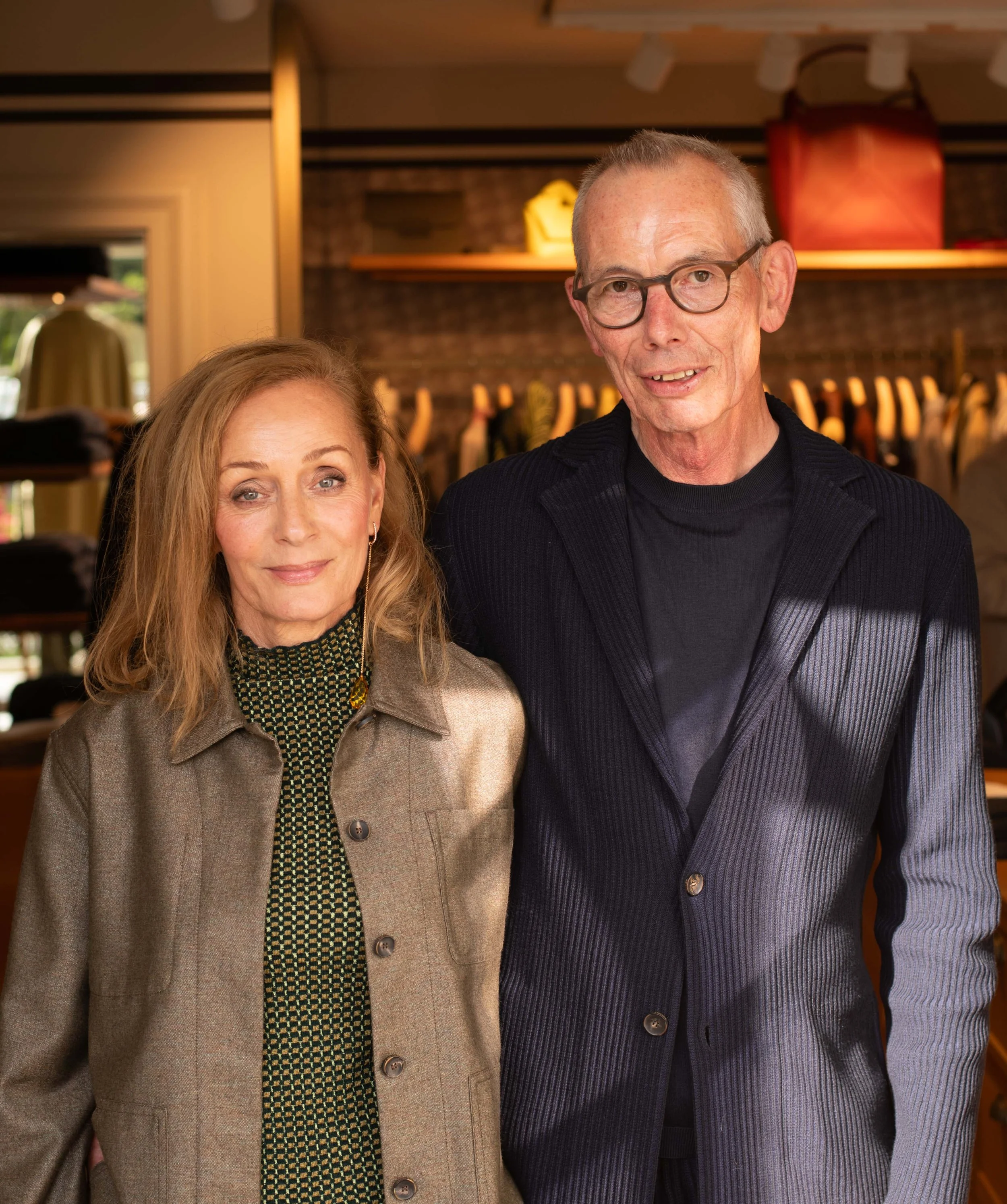 Ein älteres Paar steht in einem Laden, im Hintergrund hängen Kleidung und Taschen. Beide lächeln leicht in die Kamera.