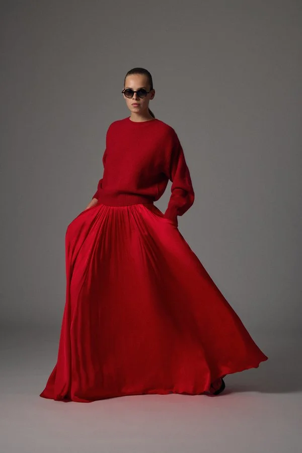 Frau in rotem Outfit, die eine lange, plissierte Rock trägt, vor einem grauen Hintergrund.