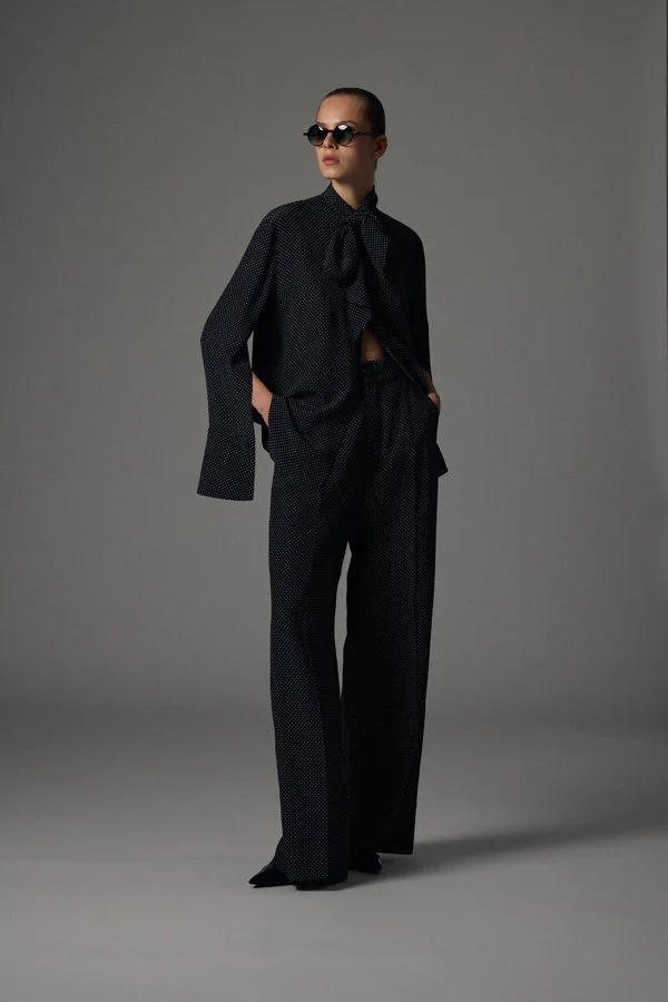 Frau trägt schwarze, gepunktete Anzughose und Bluse mit Schleife, Sonnenbrille, sitzt mit den Händen in den Taschen, vor grauem Hintergrund.