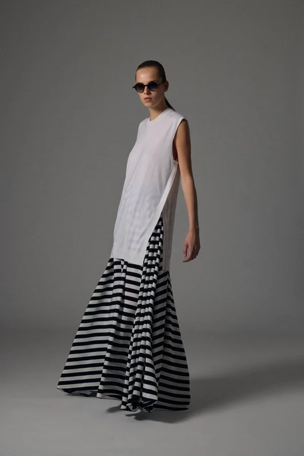 Junge Frau trägt schwarze Sonnenbrille, weiße ärmellose Tunika und schwarze-weiß gestreifte weite Hose, steht vor grauem Hintergrund.