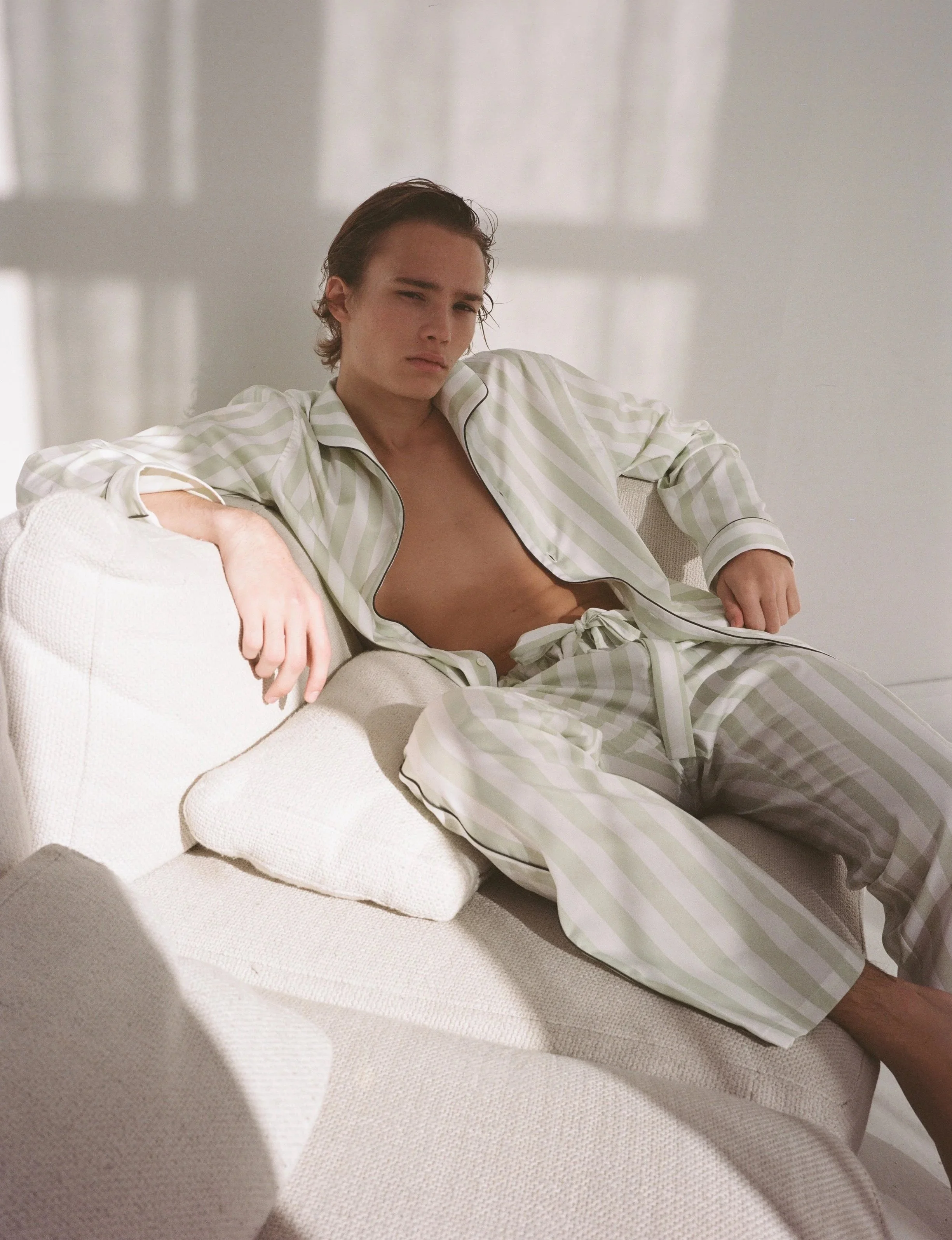 Junge Person in grüner gestreifter Pyjama sitzt auf einem weißen Sofa, schaut ernst in die Kamera, natürliche Beleuchtung, minimalistisches Zimmer im Hintergrund.